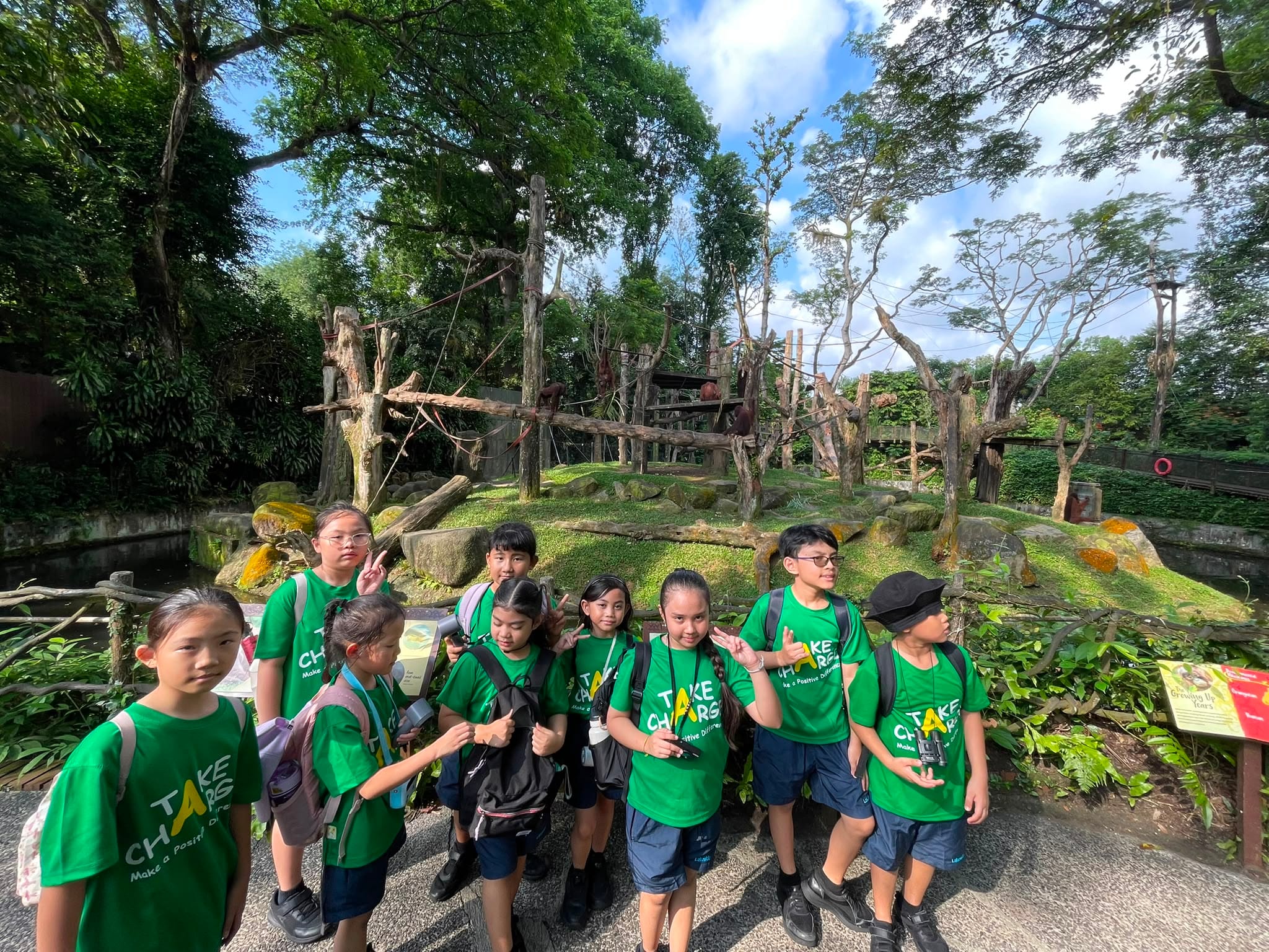 A group of students in green T-shirts standing in front of a chimpanzee enclosure with trees, rocks, and wooden structures in the background.