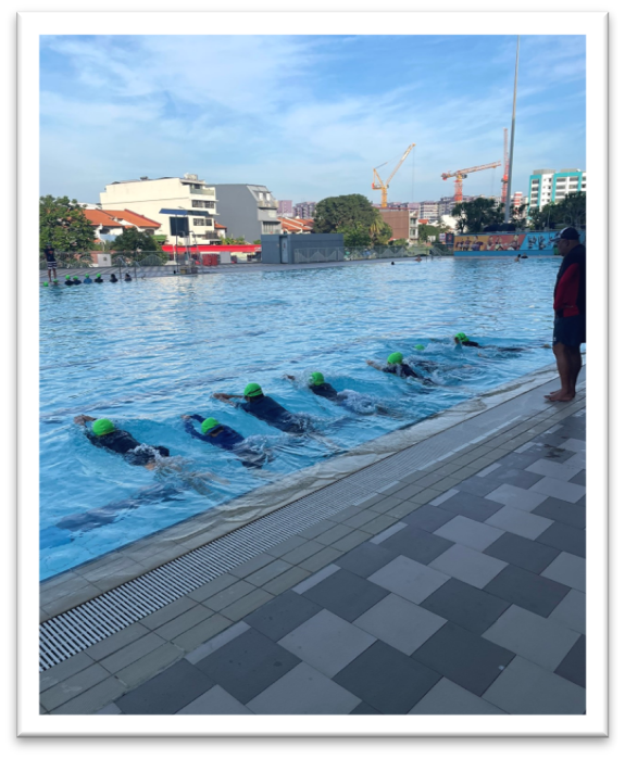 Several swimmers in green caps swim in a row in an outdoor pool while an instructor stands at the edge.