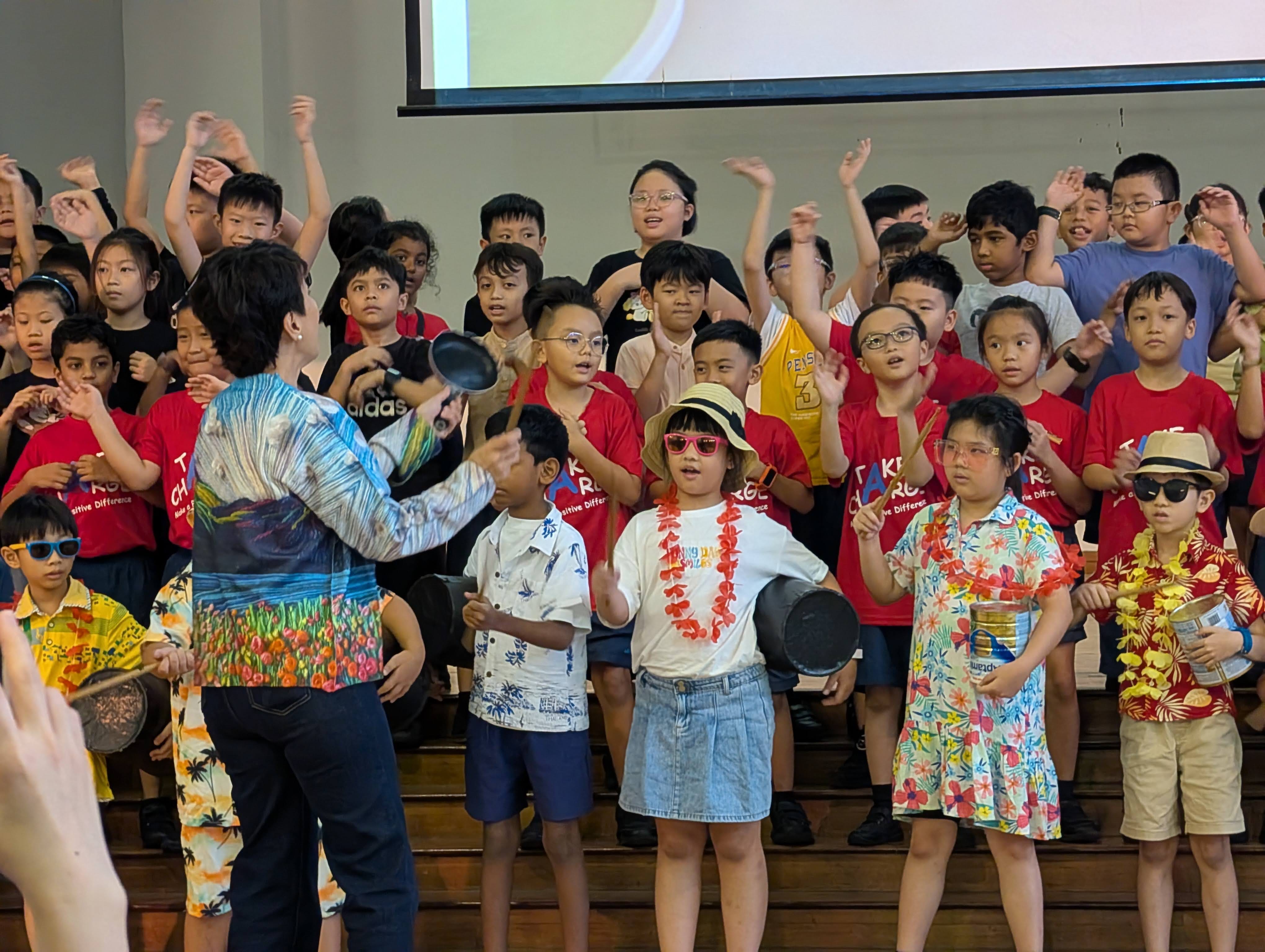Children in red shirts and tropical outfits perform on stage, holding pans, while an adult conductor leads them.