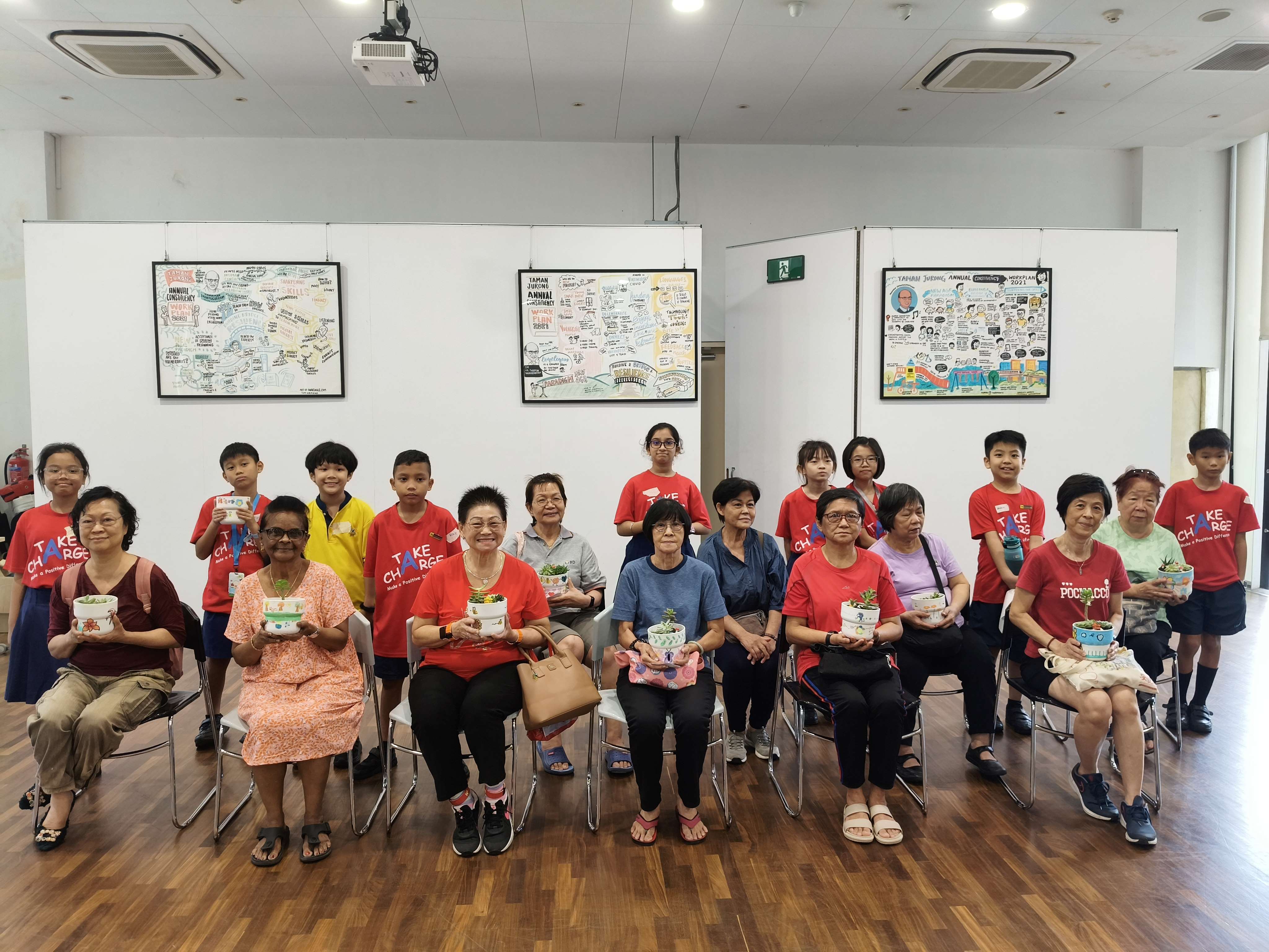 A group of adults and children in red shirts pose indoors, holding decorated plant bowls, with framed artwork on the wall.
