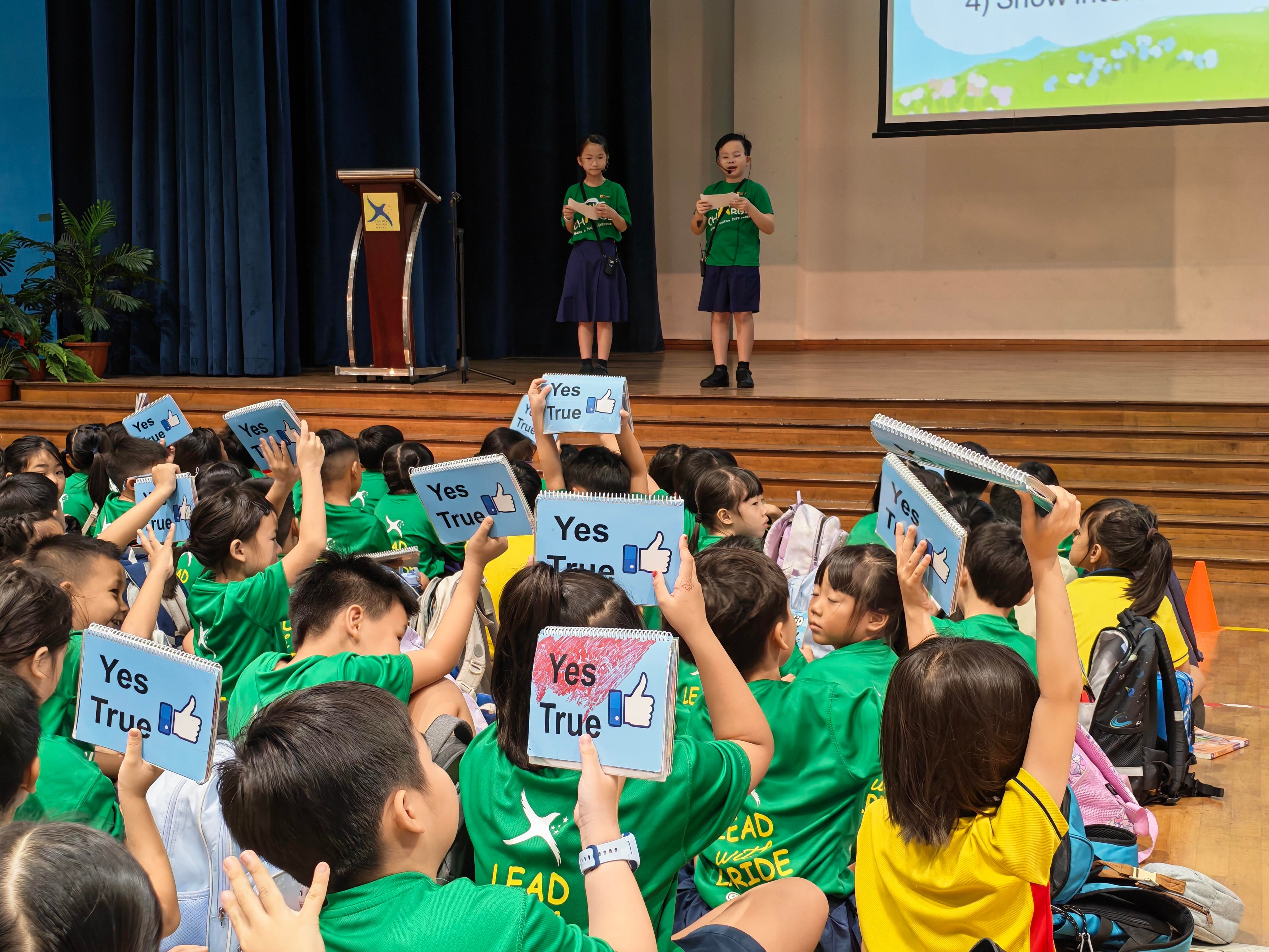 Students enthusiastically participate in a Racial Harmony Day activity by raising response cards while two student emcees facilitate the programme on stage.