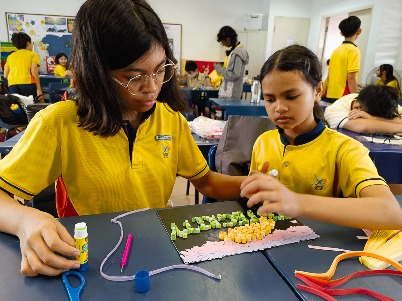 Two students in yellow uniforms work together on a craft project using small colorful pieces on a dark board.