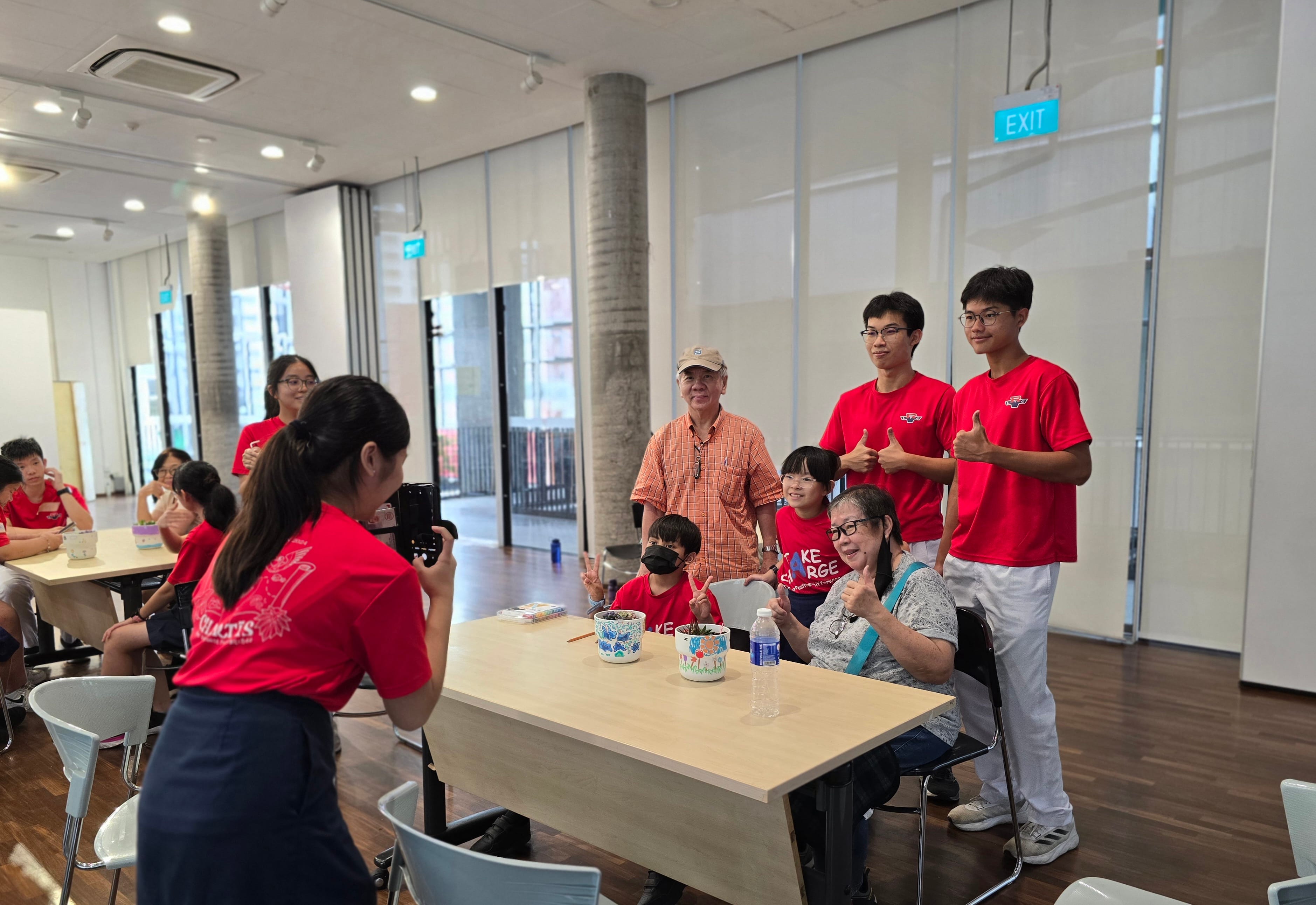 A group of people in red shirts pose with thumbs up while a girl photographs them in a bright indoor hall.