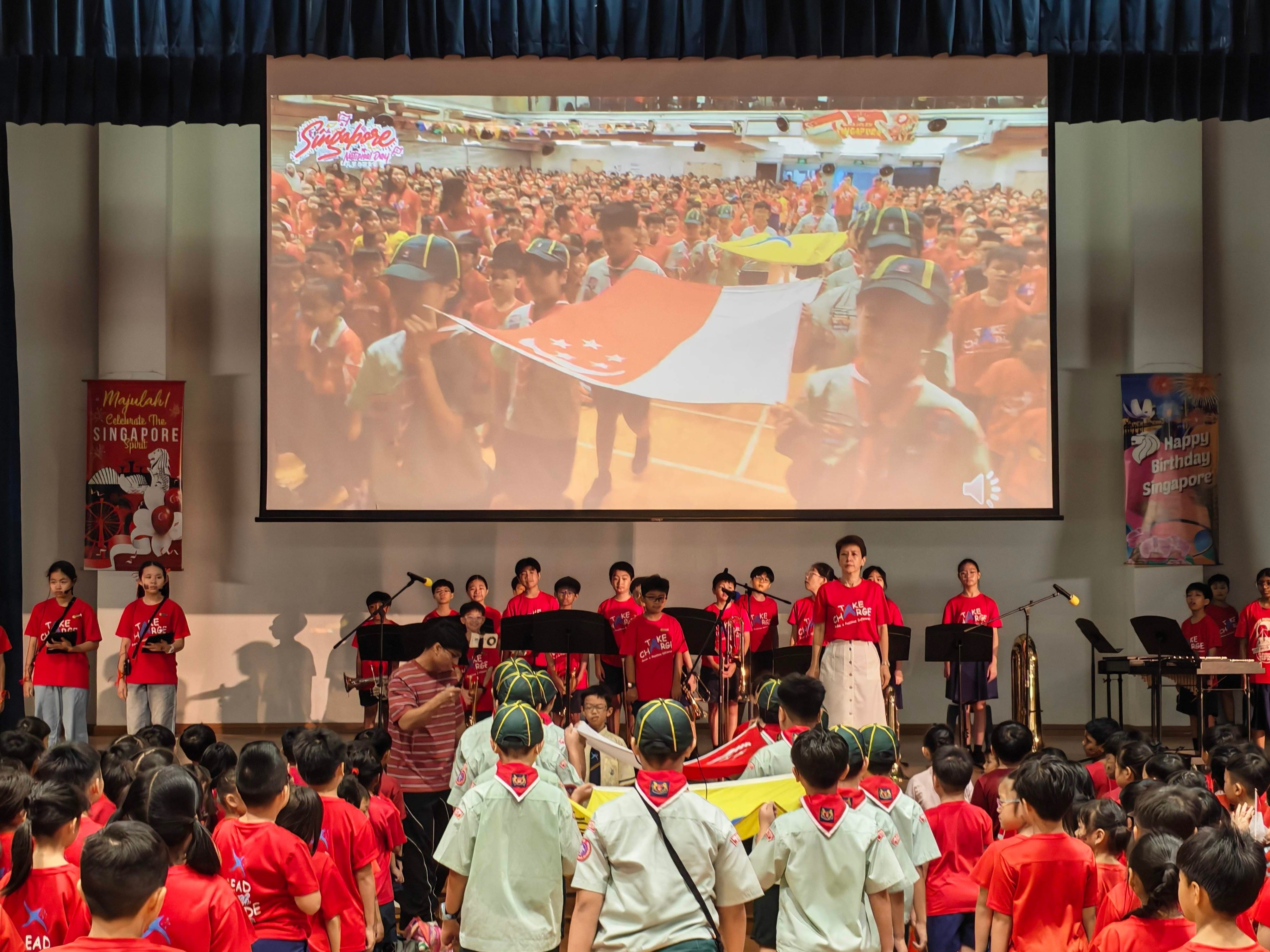 Students dressed in red gather in the school hall during the National Day Observance Ceremony, as Scouts carry the Singapore flag through the crowd and student performers stand on stage.