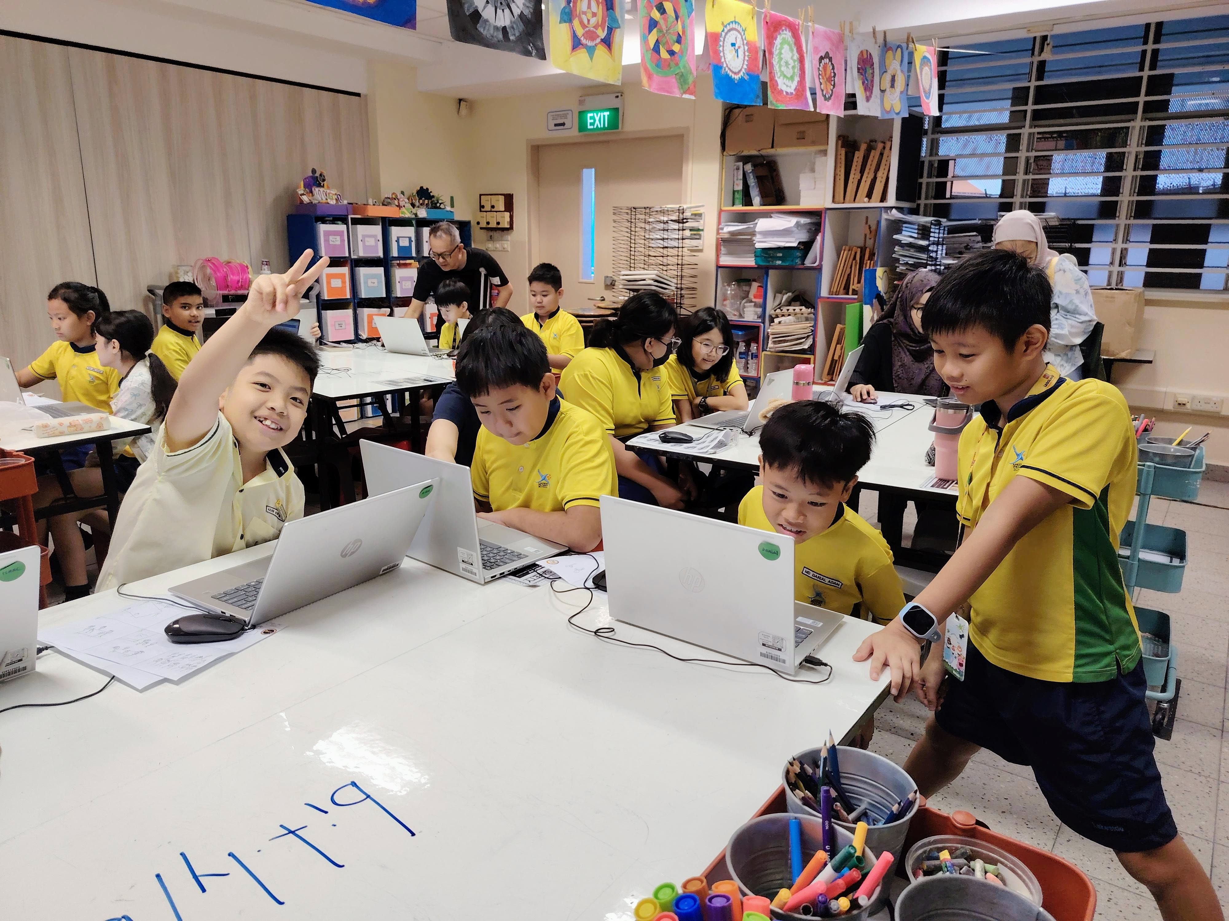 Children in yellow uniforms work on laptops in a colorful classroom; one boy smiles and holds up a peace sign.