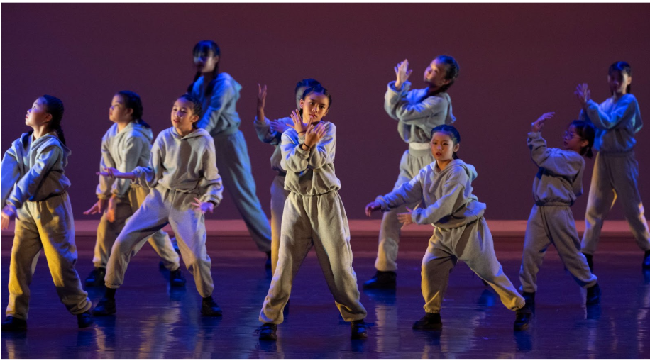 A group of young dancers in gray jumpsuits perform synchronized martial arts-style moves on a purple-lit stage.