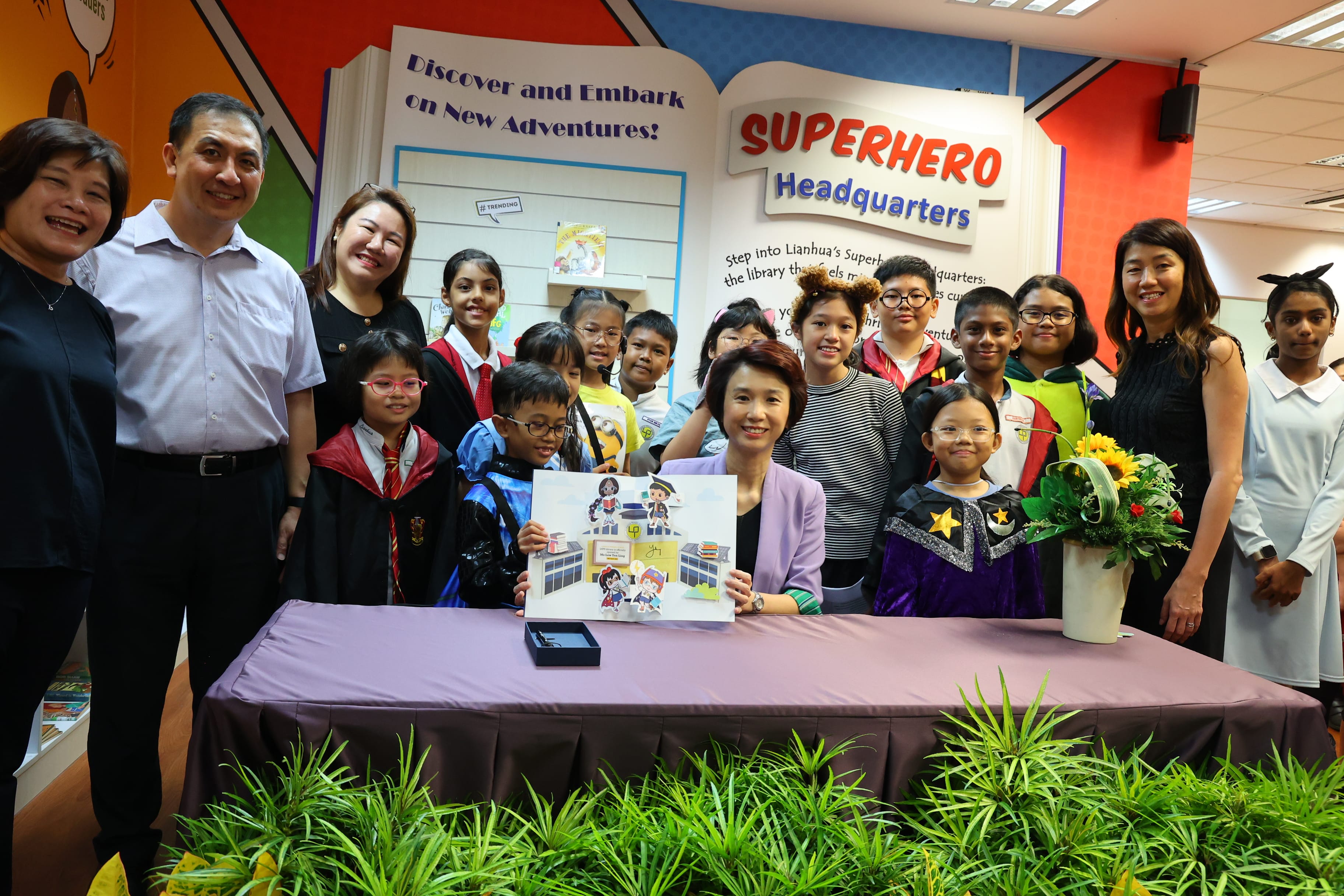 Group of adults and kids in costumes pose by a table under a Superhero Headquarters sign.