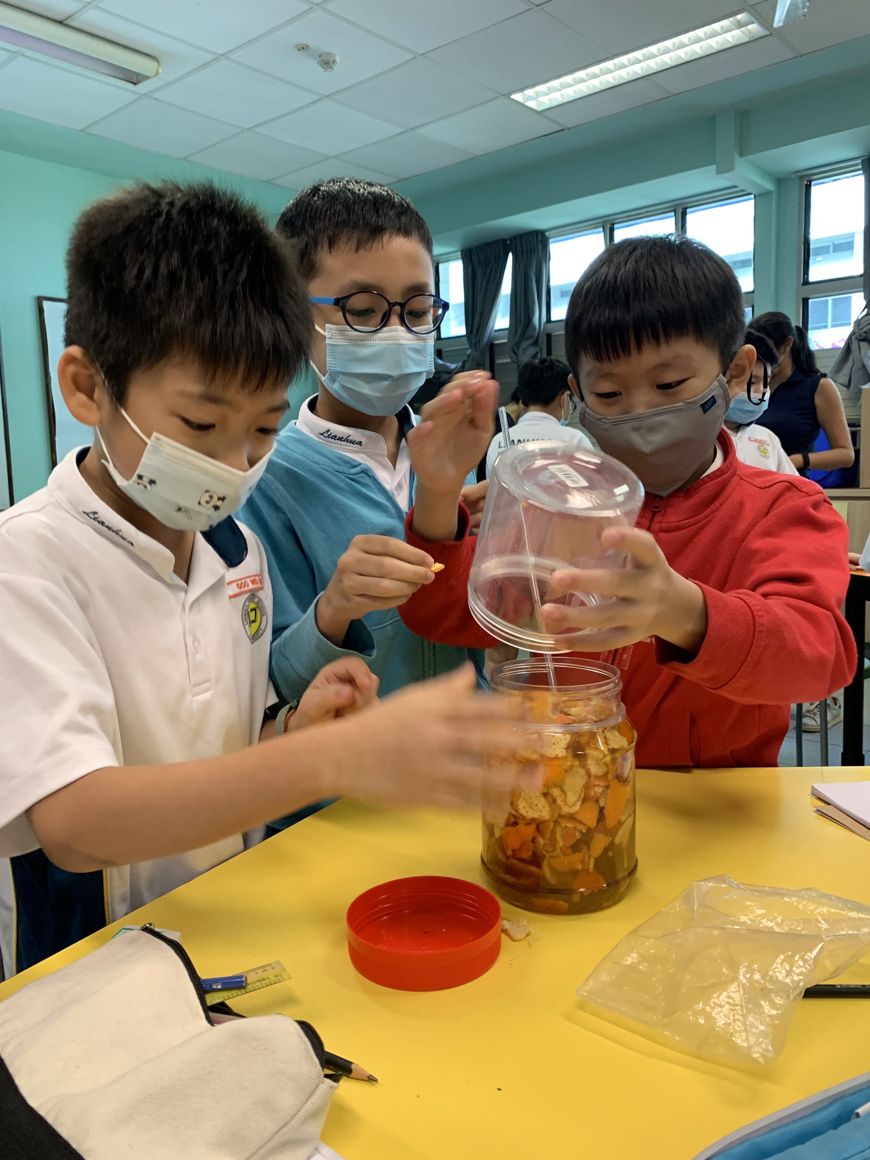 Three masked children in a classroom add food scraps into a clear jar on a yellow desk.
