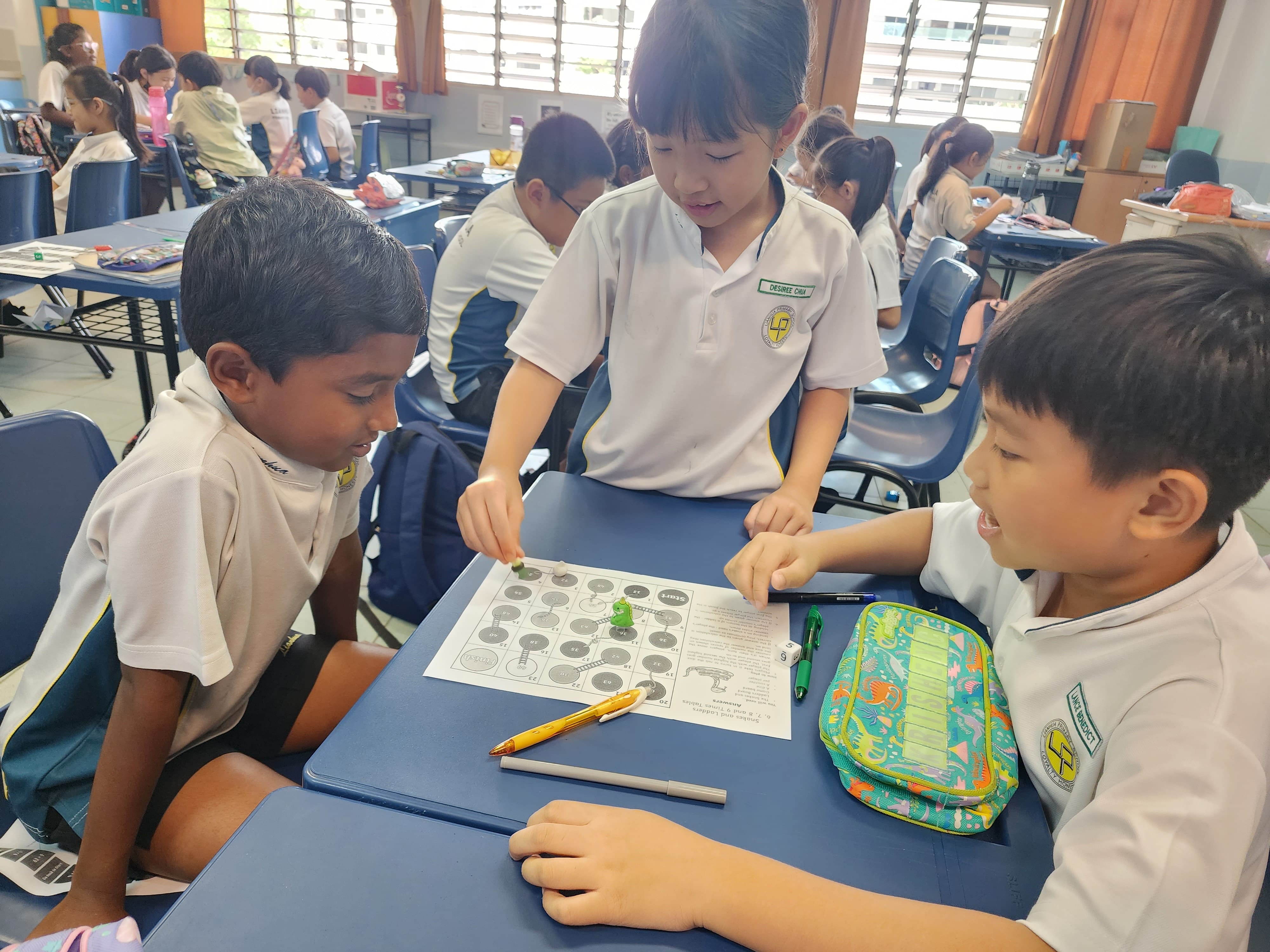 Three school children in uniforms play a snakes and ladders board game together at a blue desk in a classroom.