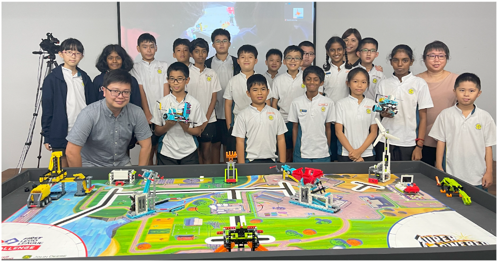 A group of children in white uniforms and an adult pose behind a FIRST LEGO League competition table with colorful LEGO builds.
