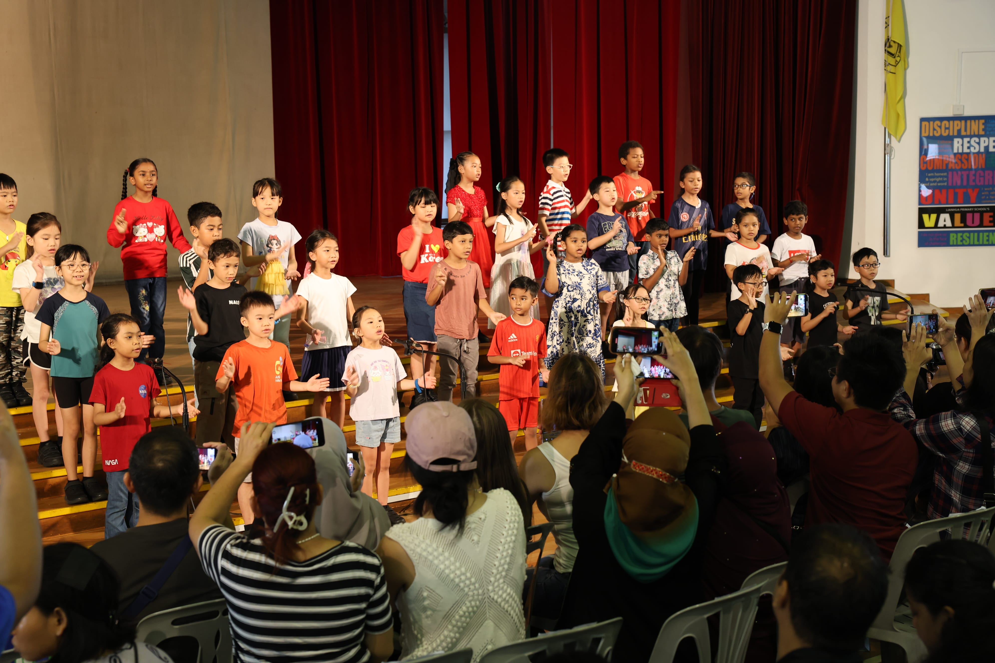 Children on stage before red curtains as audience members raise phones to take photos.