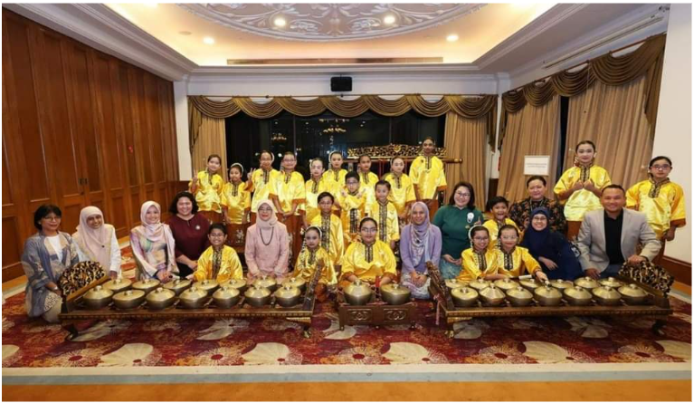 Group of people in yellow traditional outfits posing with gamelan instruments in an ornate indoor hall.
