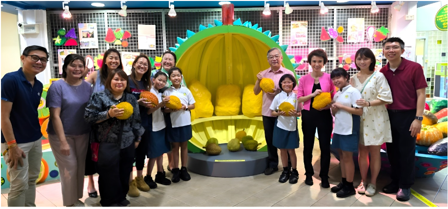 Group of adults and children posing with durian fruits in front of a large durian-shaped display at an indoor exhibit.