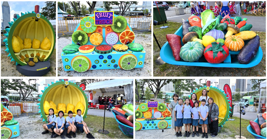 Children posing beside colorful oversized fruit and vegetable sculptures at an outdoor event with a Fruit Truck display.