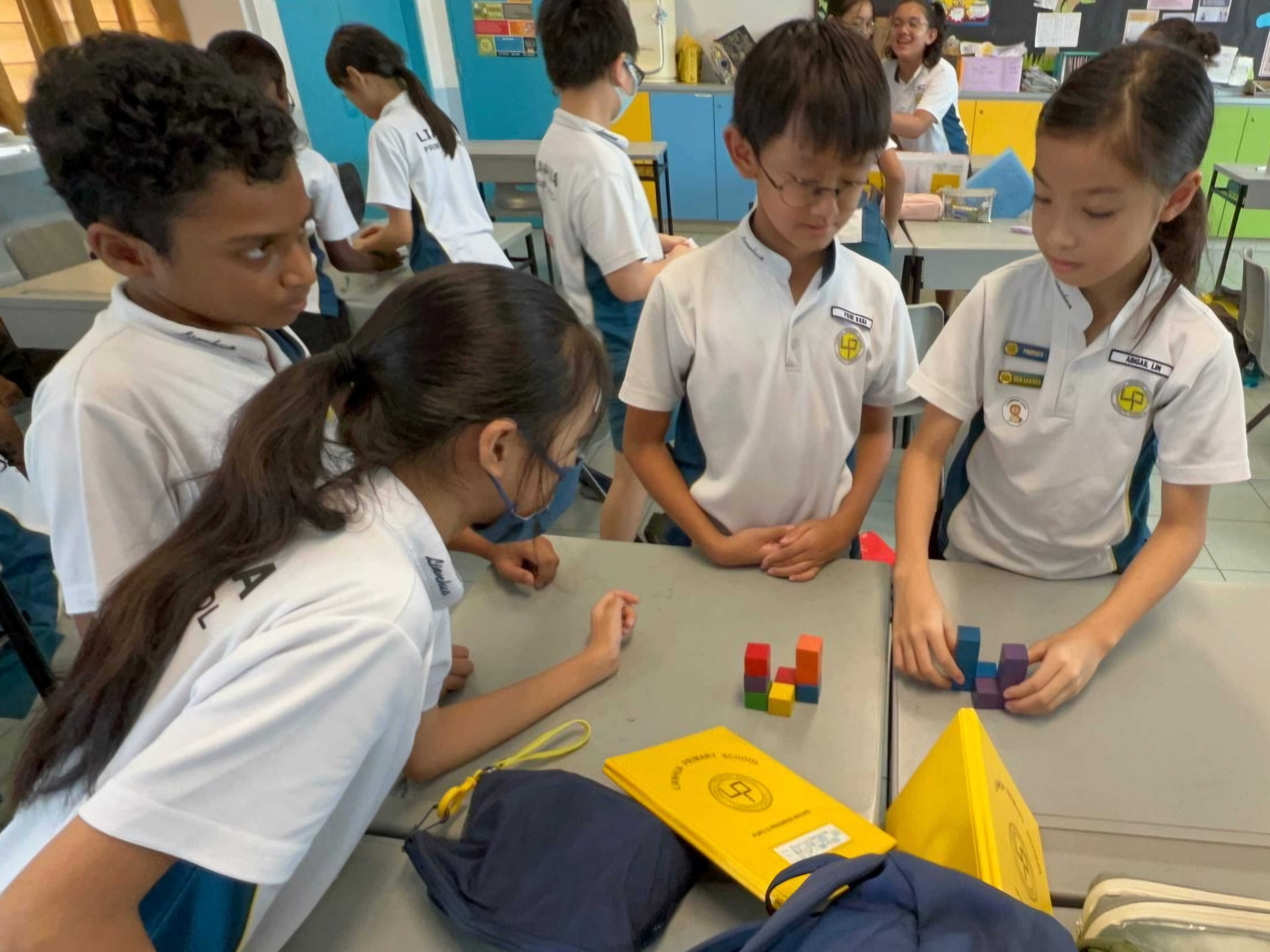 Four school children in white uniforms gather around a desk, building small colorful wooden block structures.