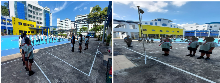 Students in school uniforms stand in formation on an outdoor courtyard near colorful blue and yellow school buildings.