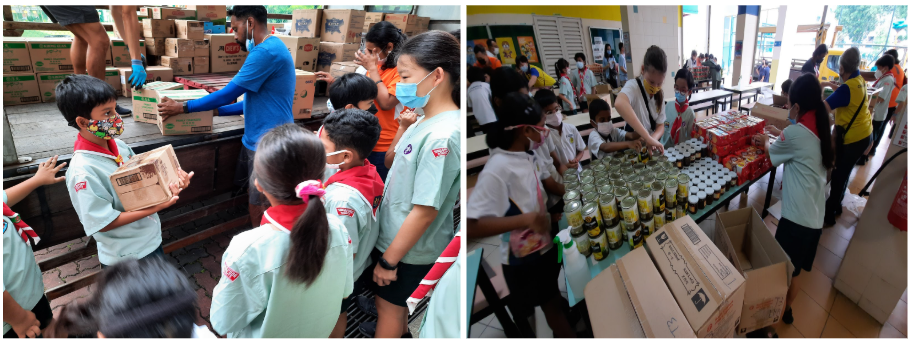 Children in uniforms and masks unload cardboard boxes from a truck and sort canned goods on tables indoors.