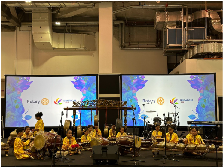 Children in yellow outfits perform traditional percussion instruments on stage at a Rotary Singapore 2024 event.