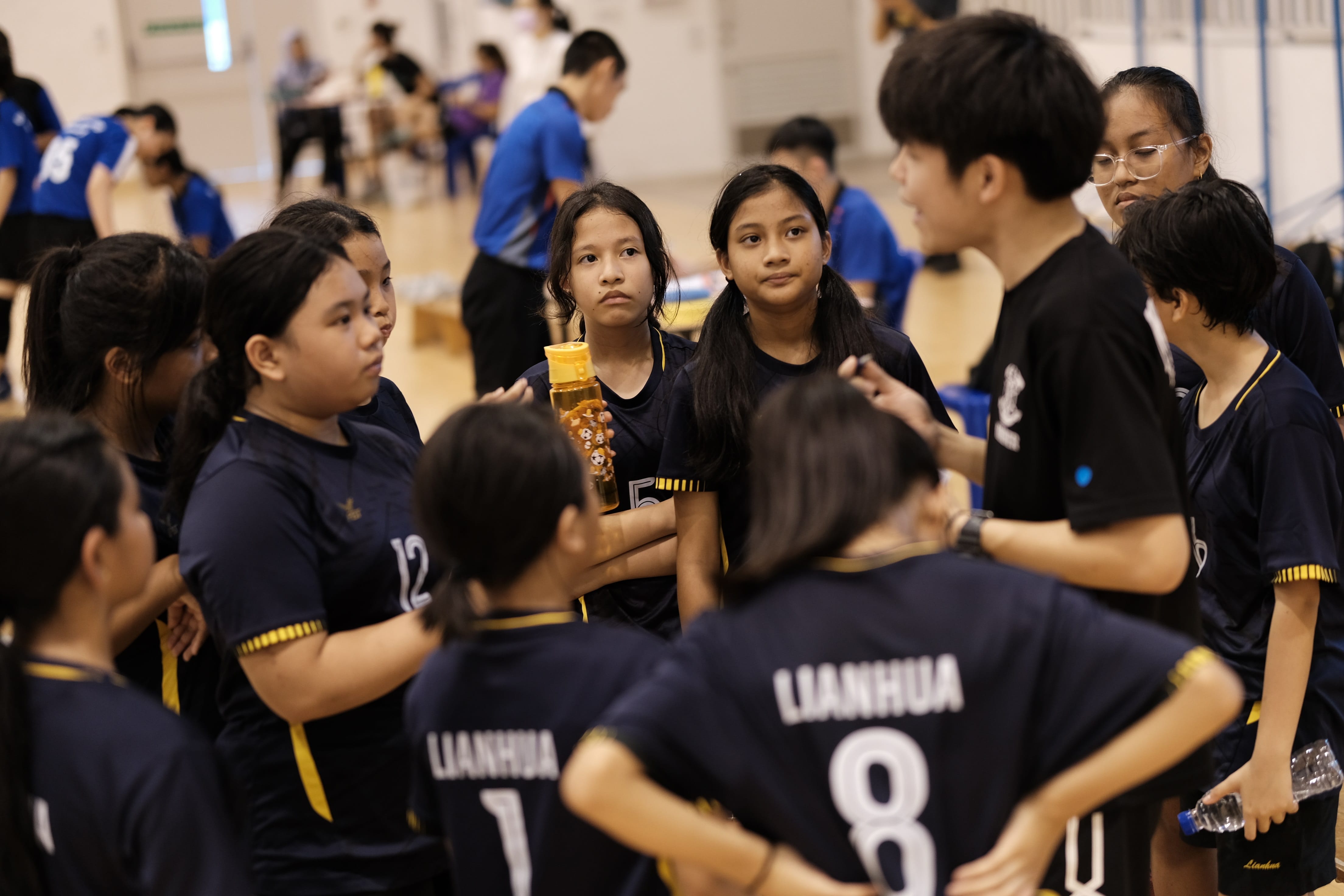 Young girls in dark navy Lianhua jerseys huddle together during a team talk at an indoor sports event.