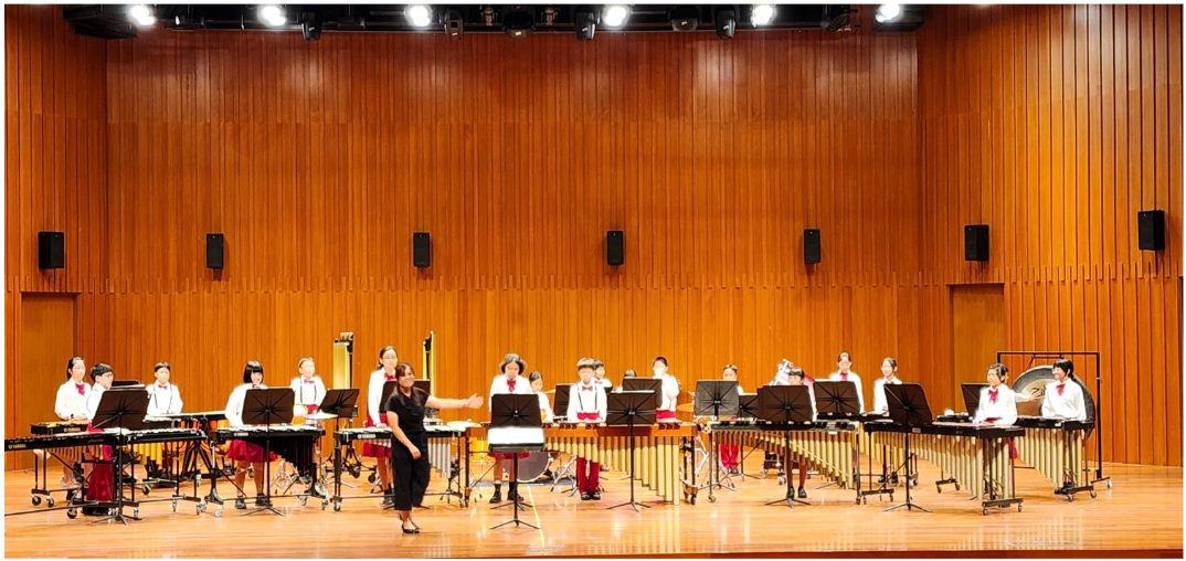 Children in white and red uniforms play xylophones and marimbas on a wooden stage while a conductor leads them.