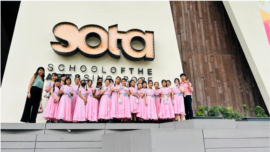 A group of children in pink dresses pose in front of the SOTA School of the Arts Singapore building.