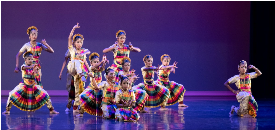A group of young classical Indian dancers in colorful striped skirts and gold headpieces perform on a lit stage.