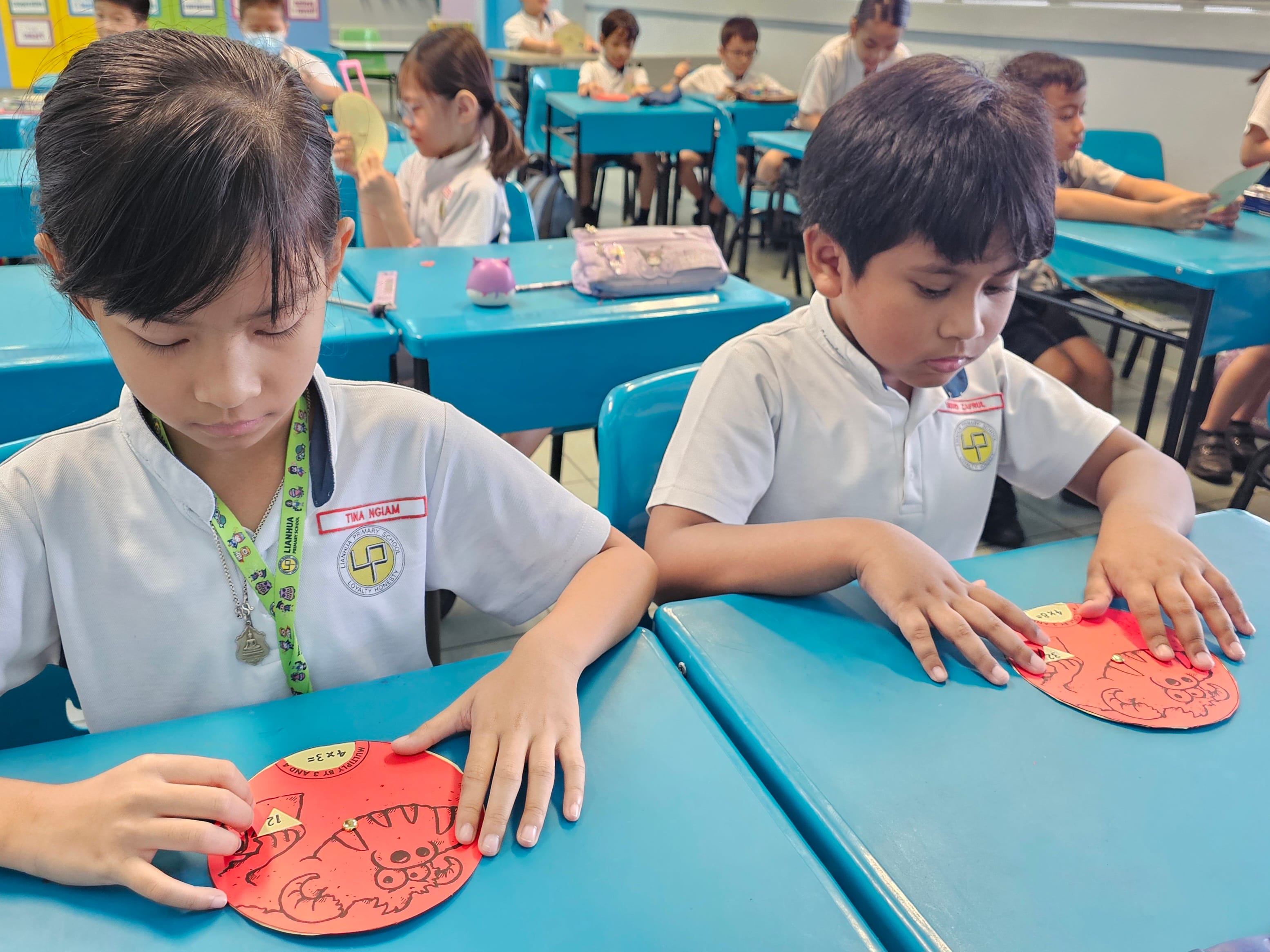 Two uniformed primary school students work on red circular multiplication craft activities at blue desks in a classroom.