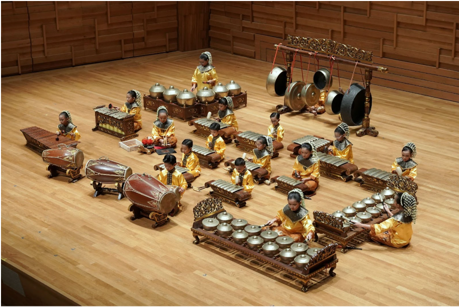 Children in gold costumes sit on a wooden stage playing traditional gamelan instruments including gongs and xylophones.