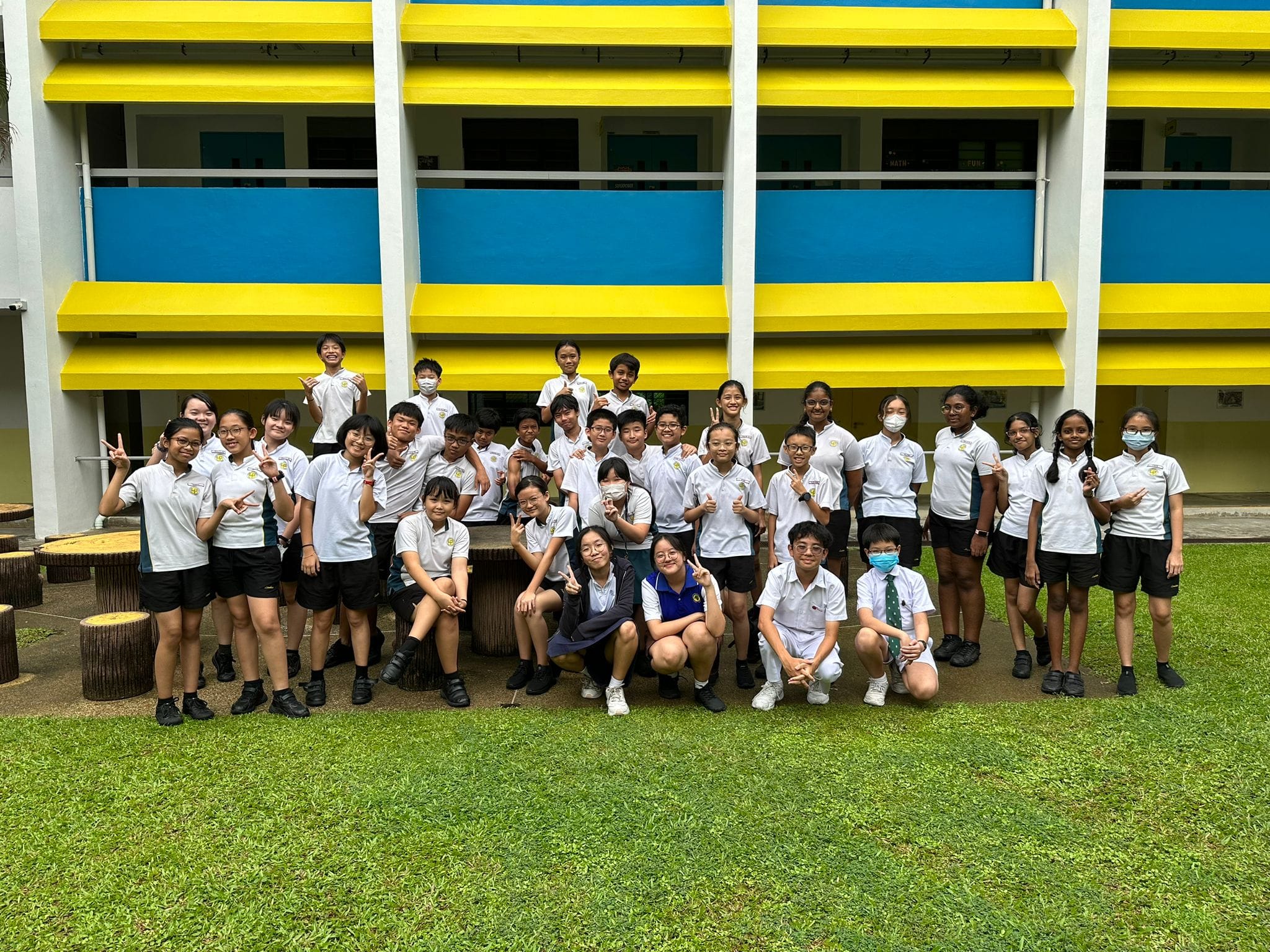 A large group of students in white uniforms posing outdoors in front of a yellow and blue school building.