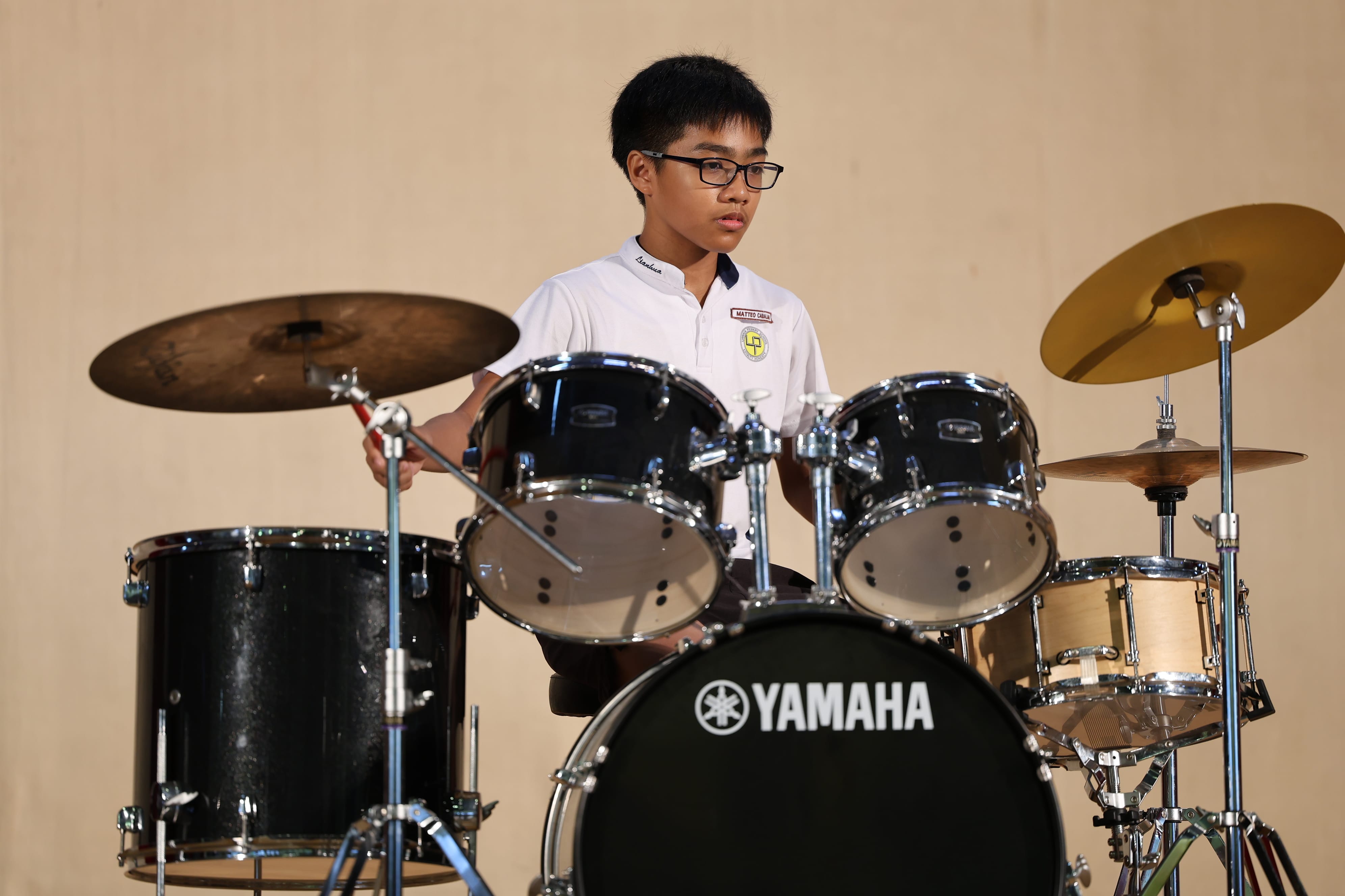 A young boy with glasses in a white shirt plays a Yamaha drum kit with cymbals on a stage.