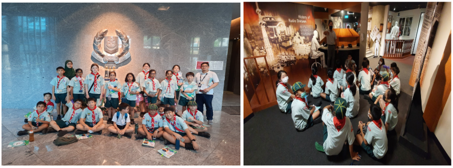 A group of young scouts in light blue uniforms and red scarves pose in a museum lobby, then sit on the floor at an exhibit.