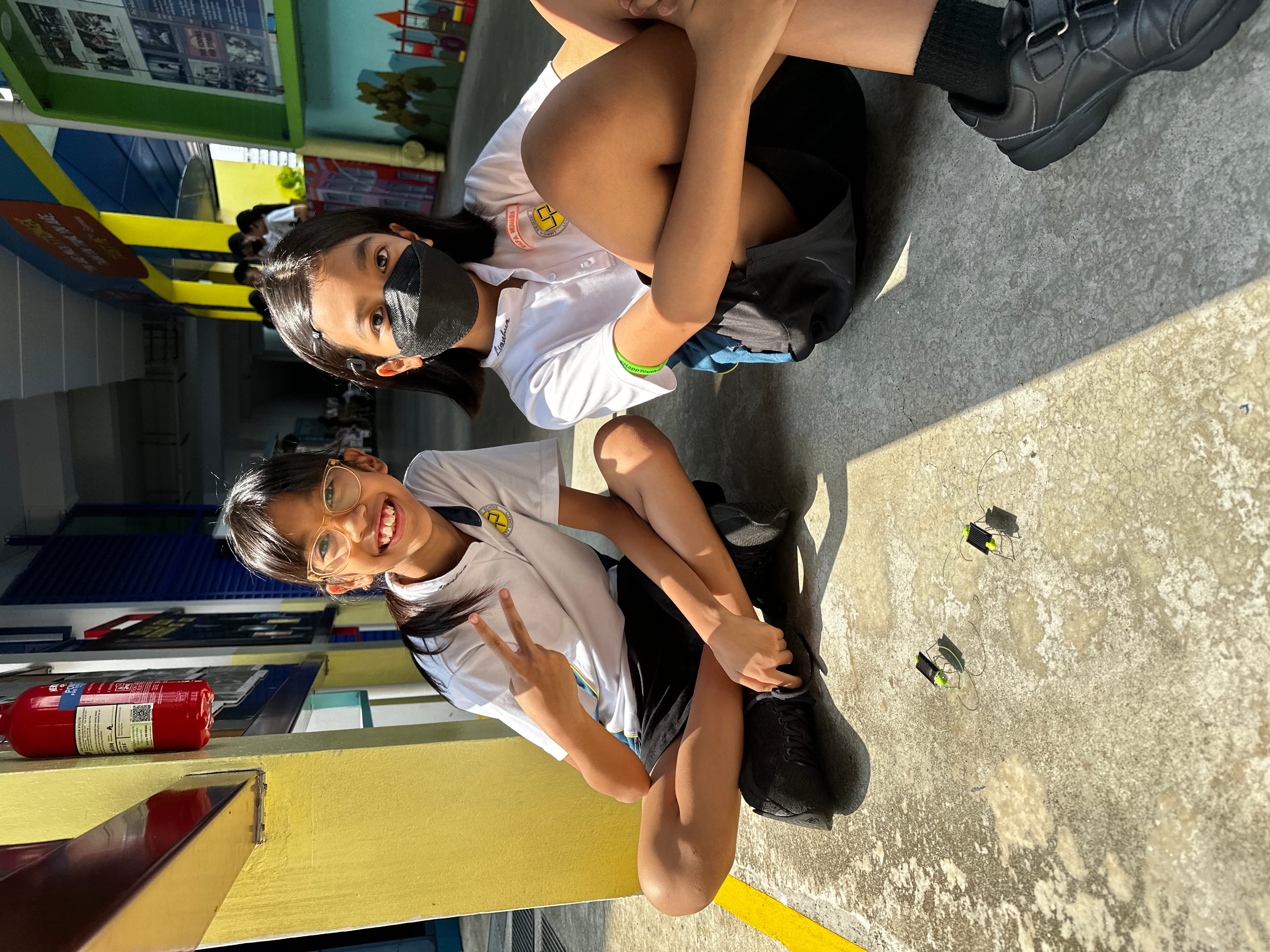 Two young schoolgirls in white uniforms sit on a corridor floor, smiling, with small electronic components in front of them.