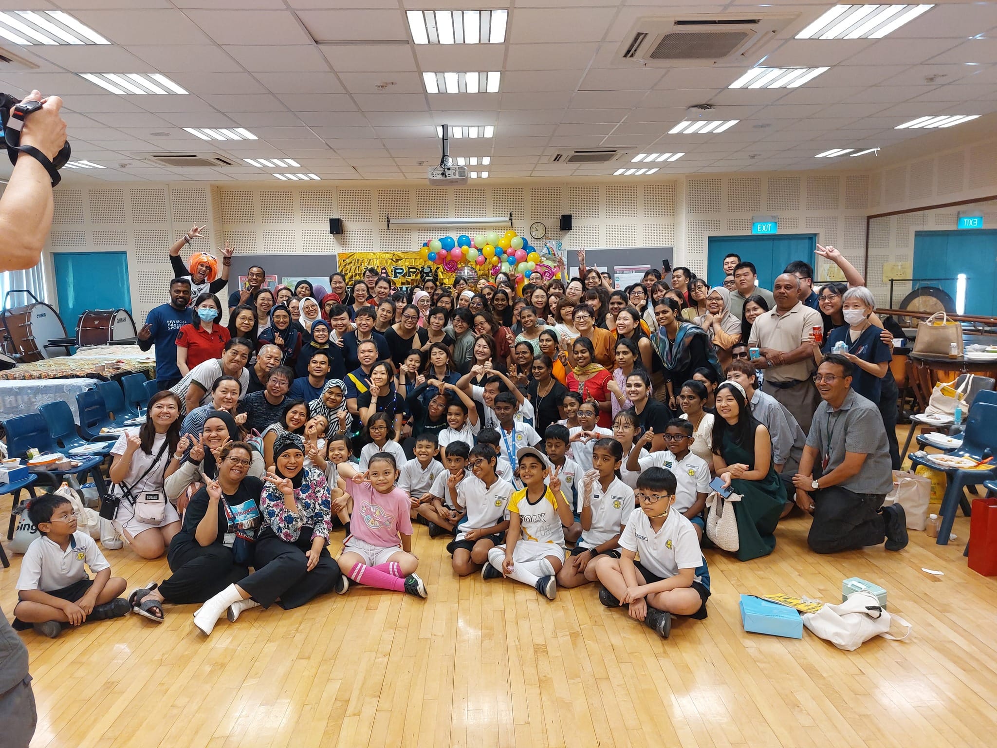 Large diverse group of adults and children posing together in a school hall with colorful balloons in the background.