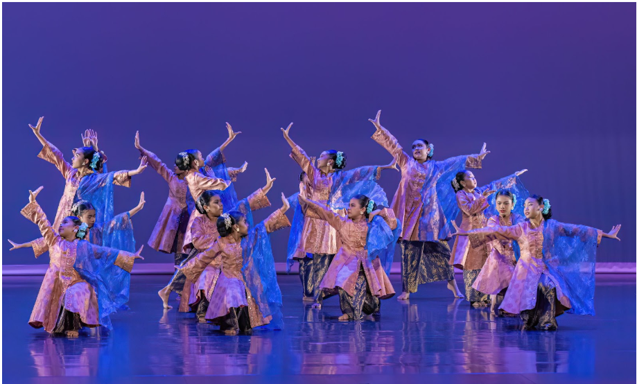 A group of dancers in blue and gold traditional costumes perform on a purple-lit stage with arms raised.
