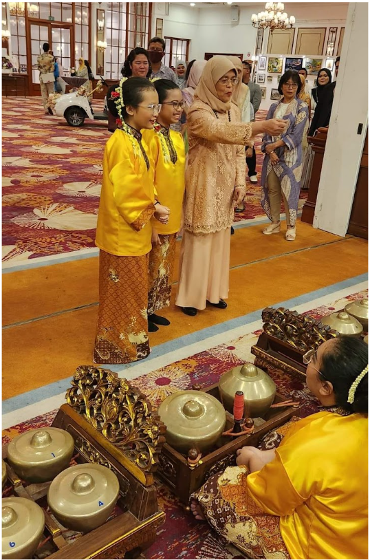 Children in yellow traditional outfits stand near gamelan instruments while an older woman in beige lace speaks to them indoors.