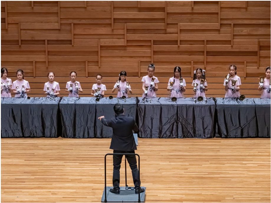 A conductor in black leads a row of young girls in pink holding handbells on a wooden concert stage.