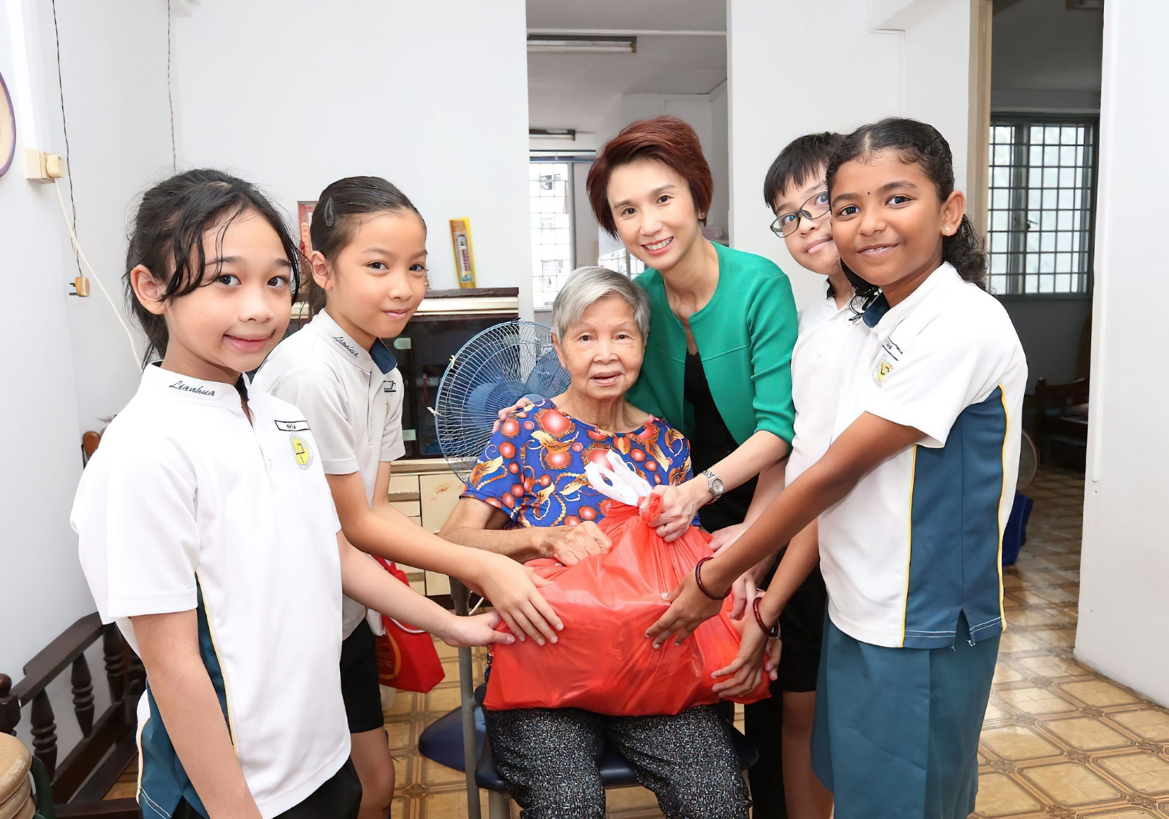 Four schoolchildren and a woman in green present a red gift bag to a smiling elderly woman indoors.