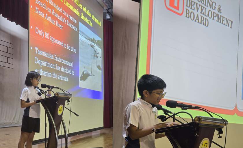 Two students in school uniforms present at a podium with projection screens showing slides behind them.