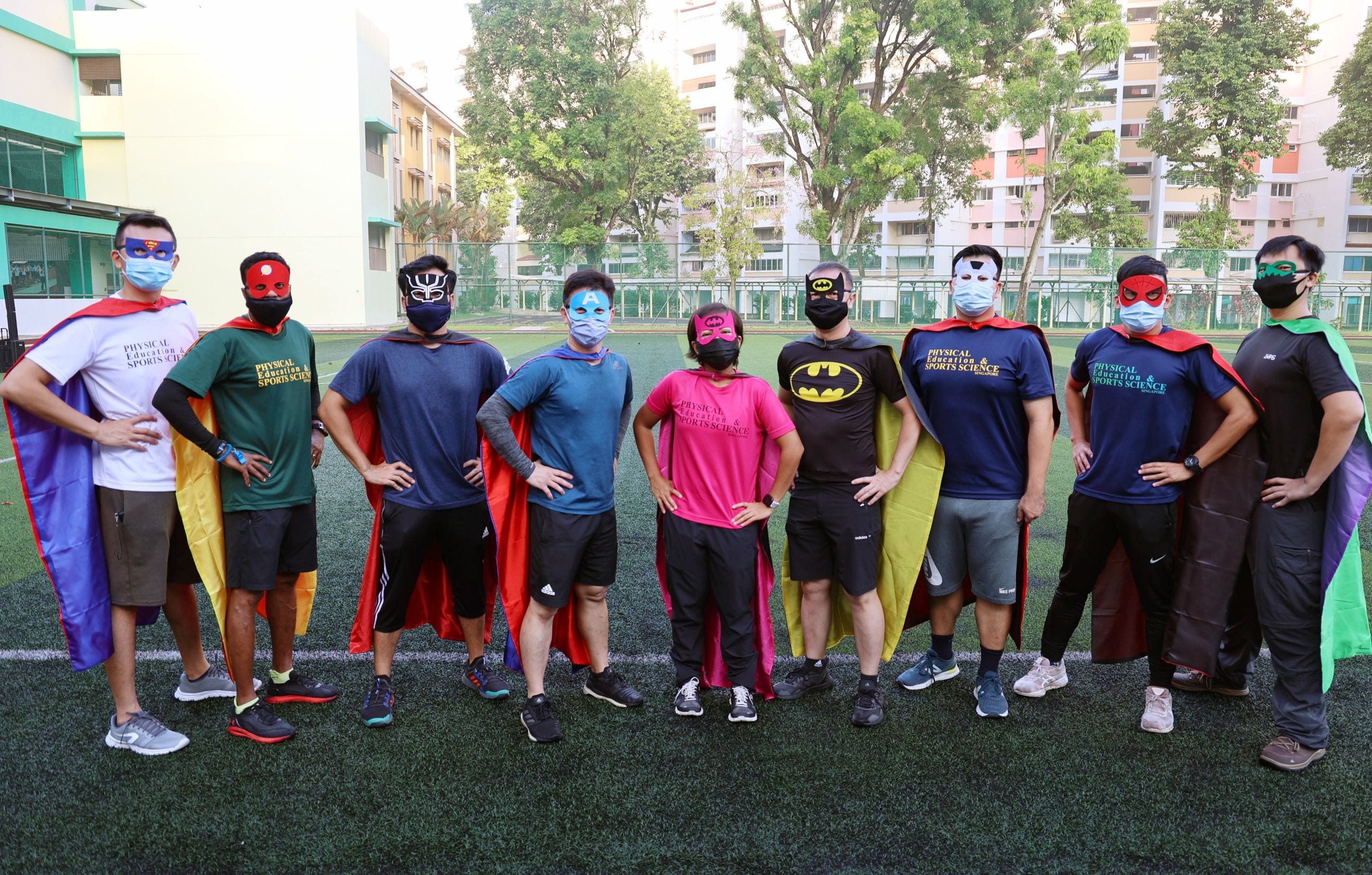 Nine people wearing superhero masks, capes, and face masks pose on a green field with buildings in the background.