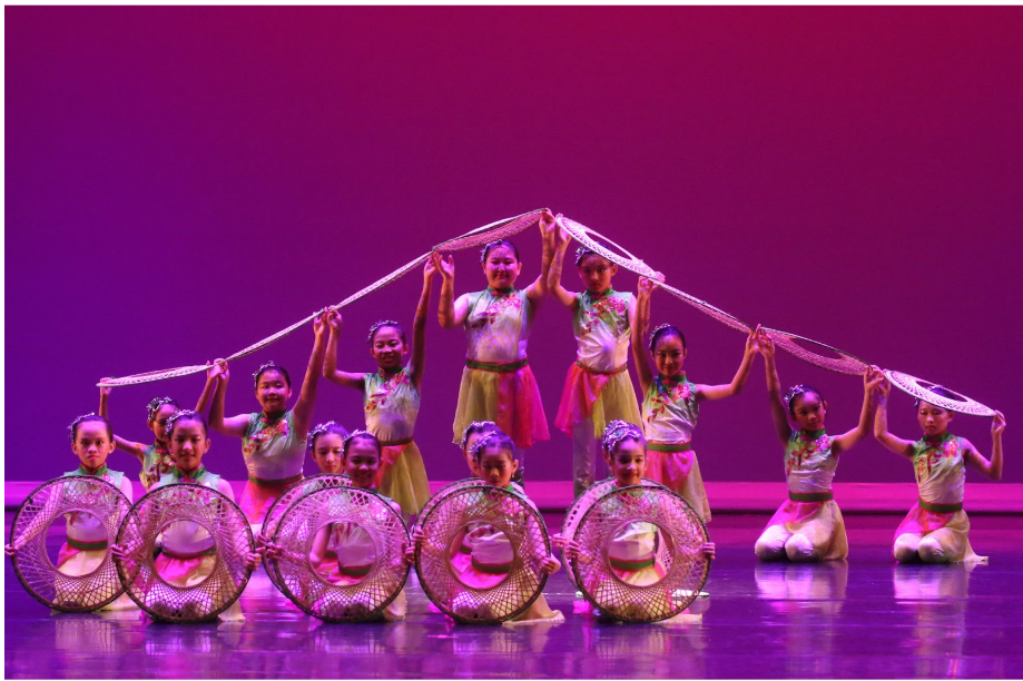 Girls in traditional Chinese costumes perform on a purple-lit stage, holding woven circular props and long ribbon garlands.