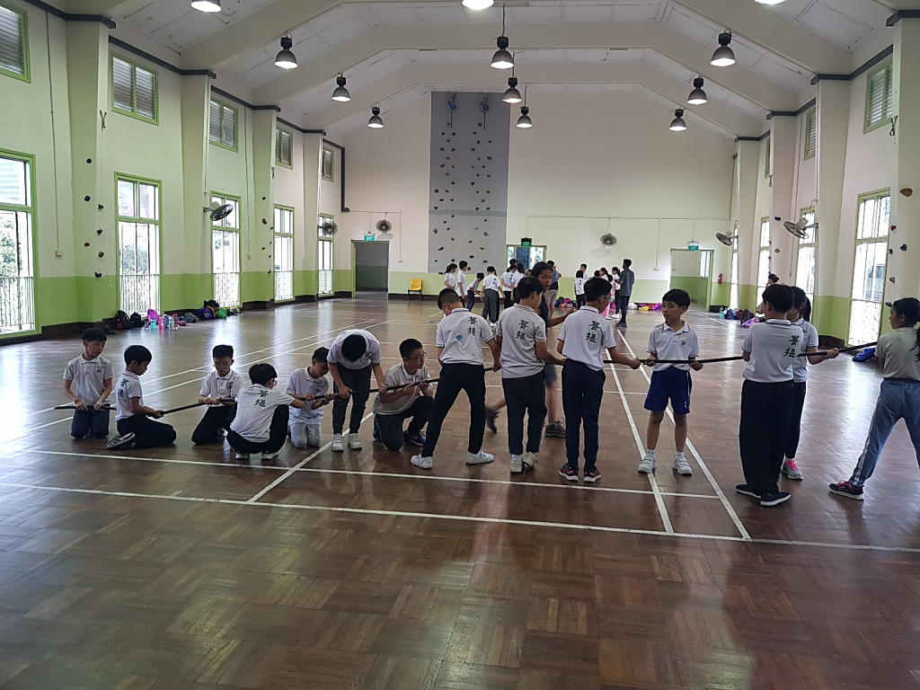 Children in white uniforms pulling a pole together in a large indoor gymnasium with a climbing wall.
