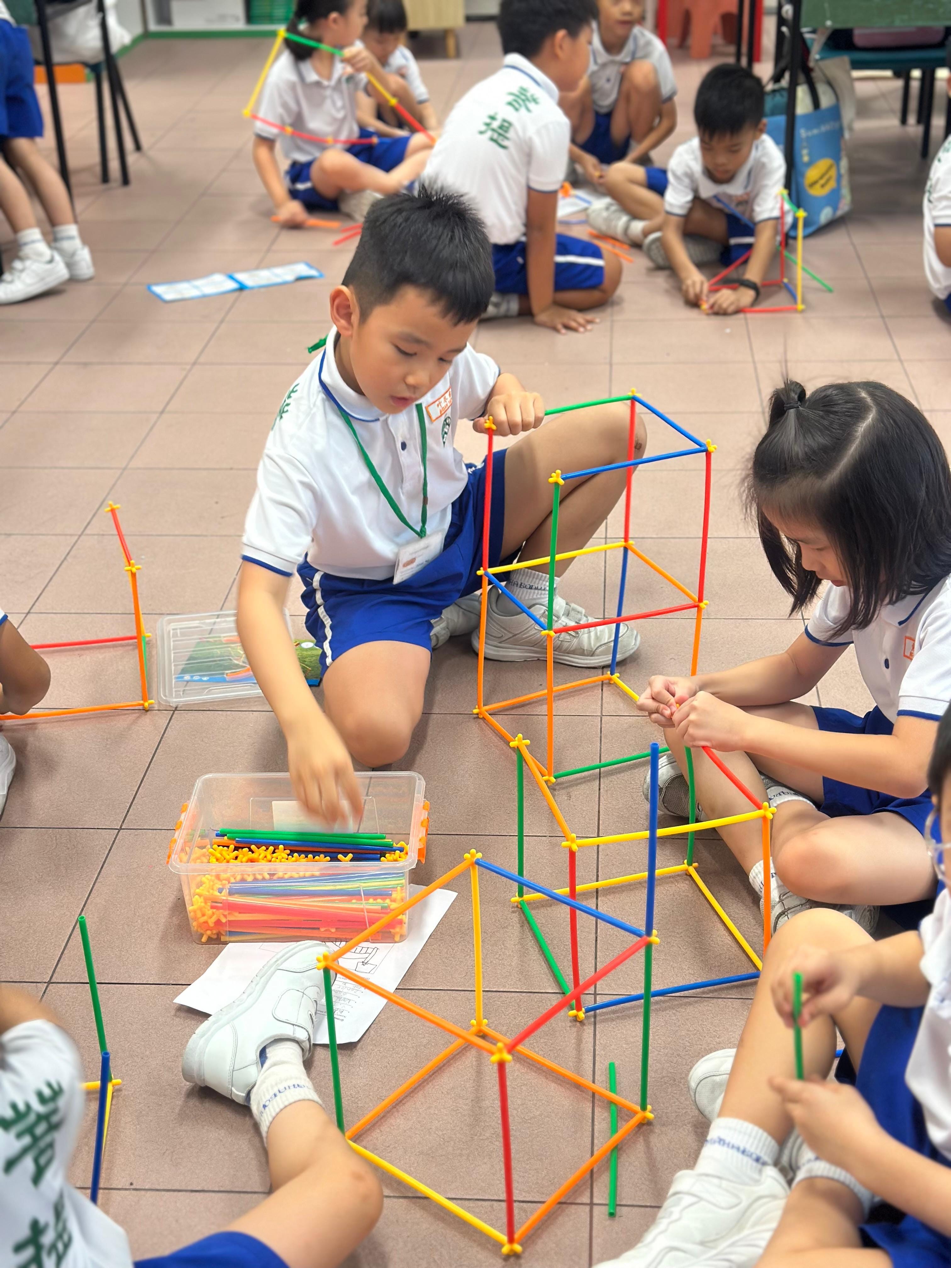 Primary 1 children in Problem Solving Challenge dress in PE attire sitting on  floor building colourful geometric structures using plastic straws and connectors to cross a river.