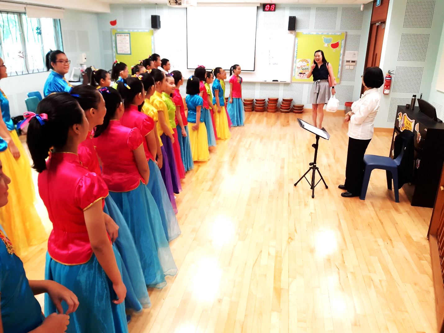 Girls in colorful traditional Chinese costumes stand in rows in a classroom, facing two adults near a piano and music stand.