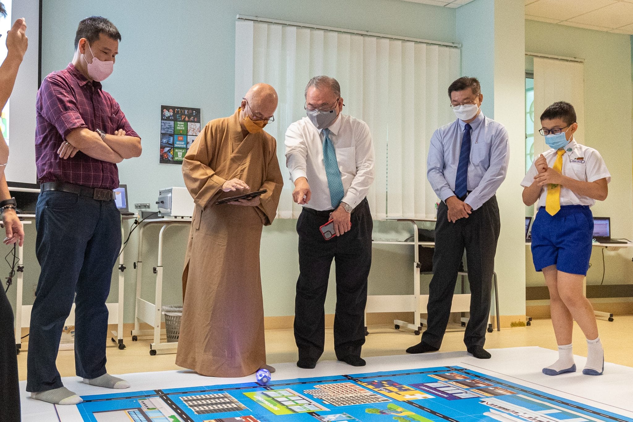 Four masked adults and a young boy in school uniform gather around a colorful floor map with a small robot on it.