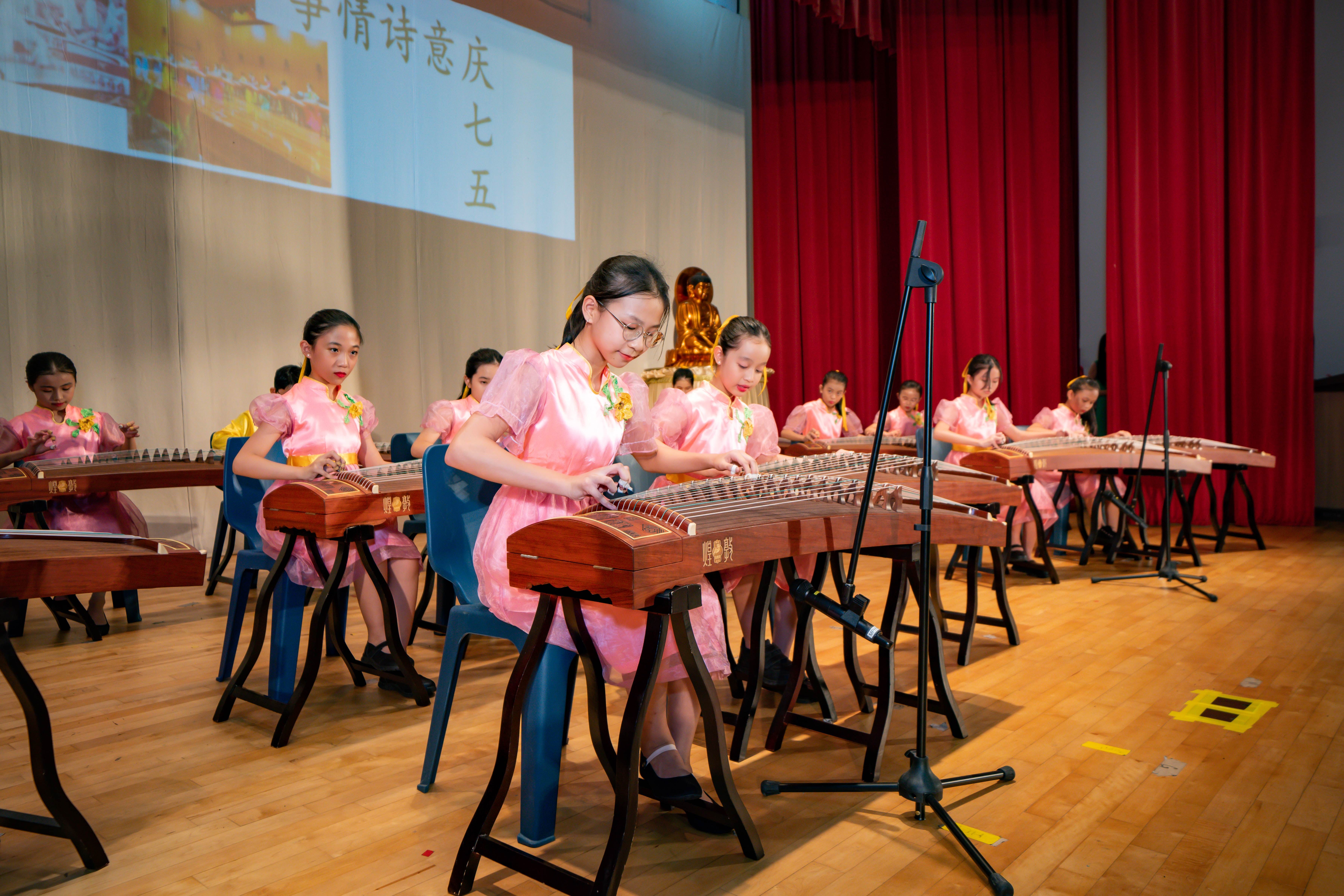 Girls in pink dresses perform on guzheng instruments on a stage with red curtains and Chinese text projected behind them.