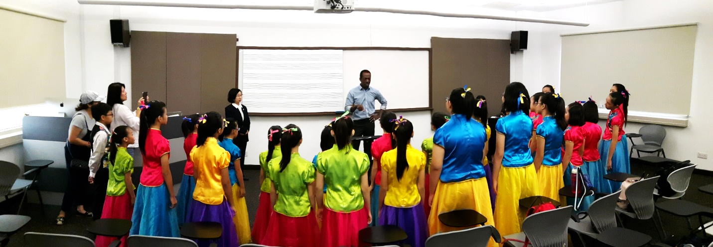 Girls in colorful traditional costumes stand in a semicircle facing a conductor in a classroom.