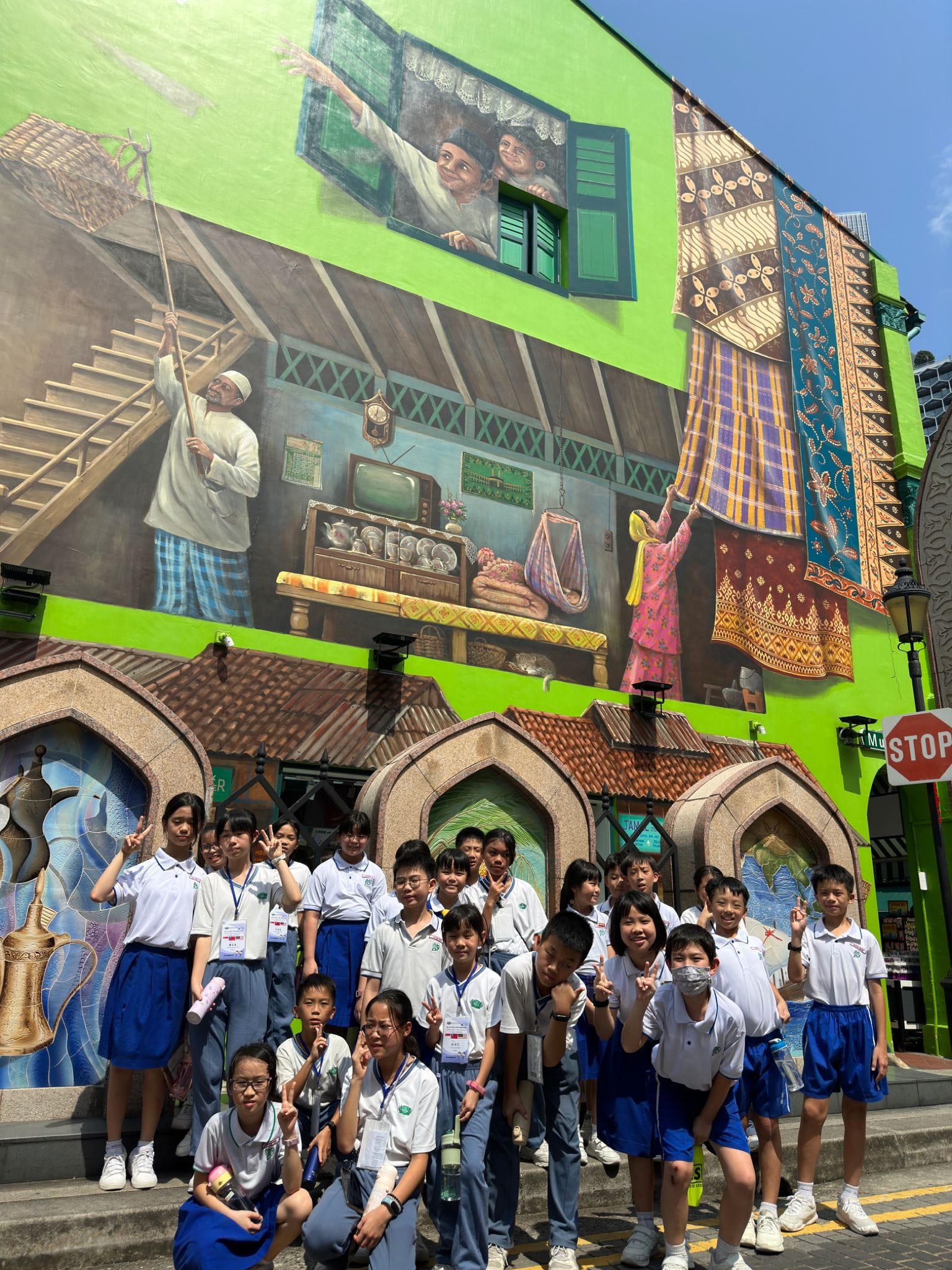 A group of schoolchildren in white and blue uniforms pose in front of a large colorful cultural street mural.
