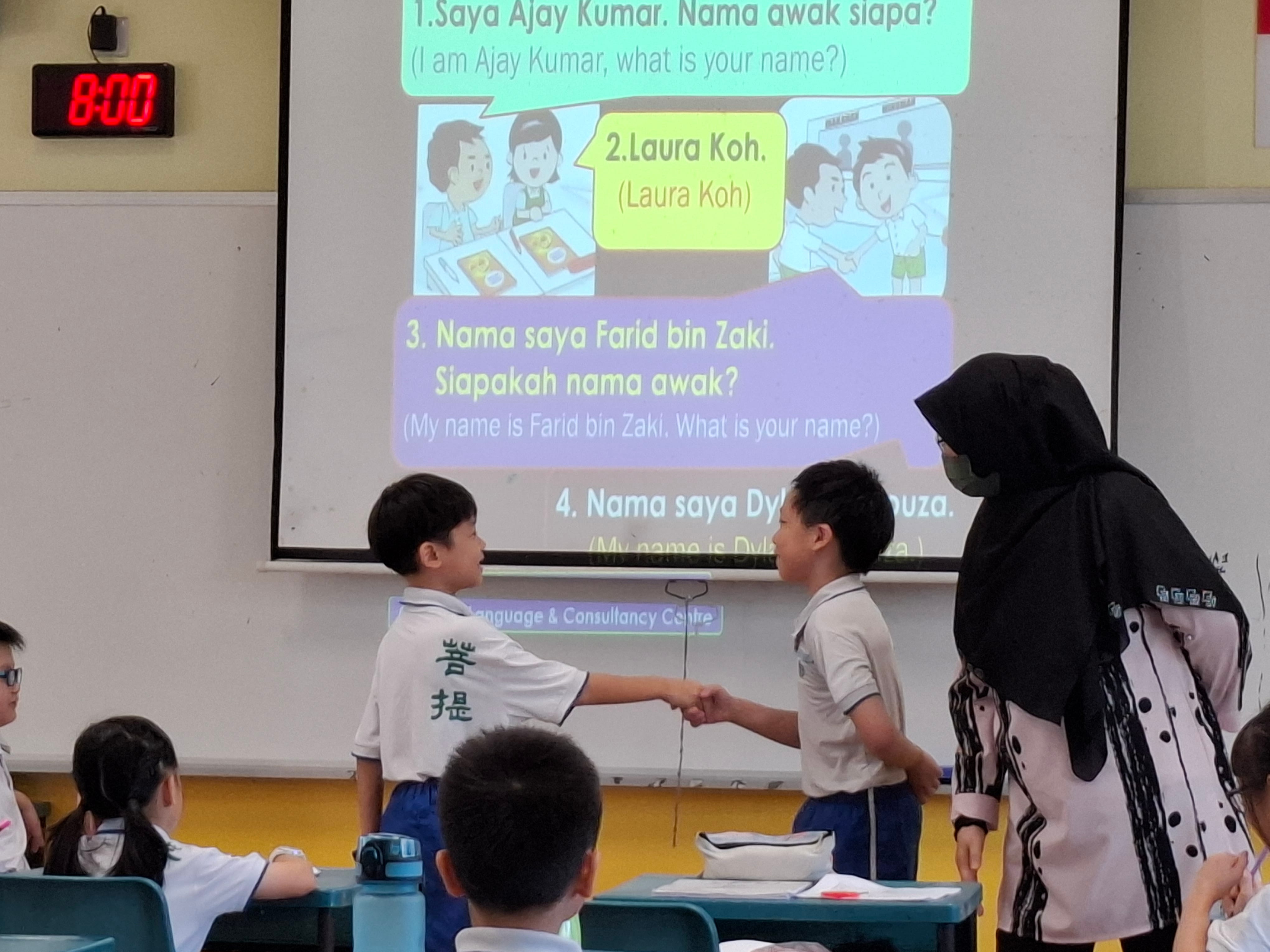 Two young students shake hands in a classroom while a teacher watches, with a Malay language lesson projected behind them.