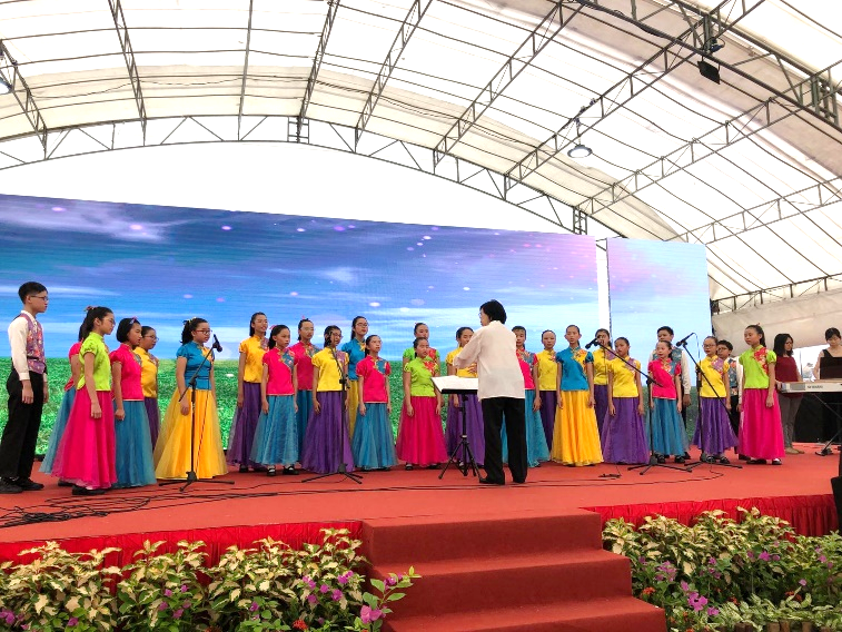 Children in colorful dresses perform on a red-carpeted stage while a conductor stands before them under a tent.