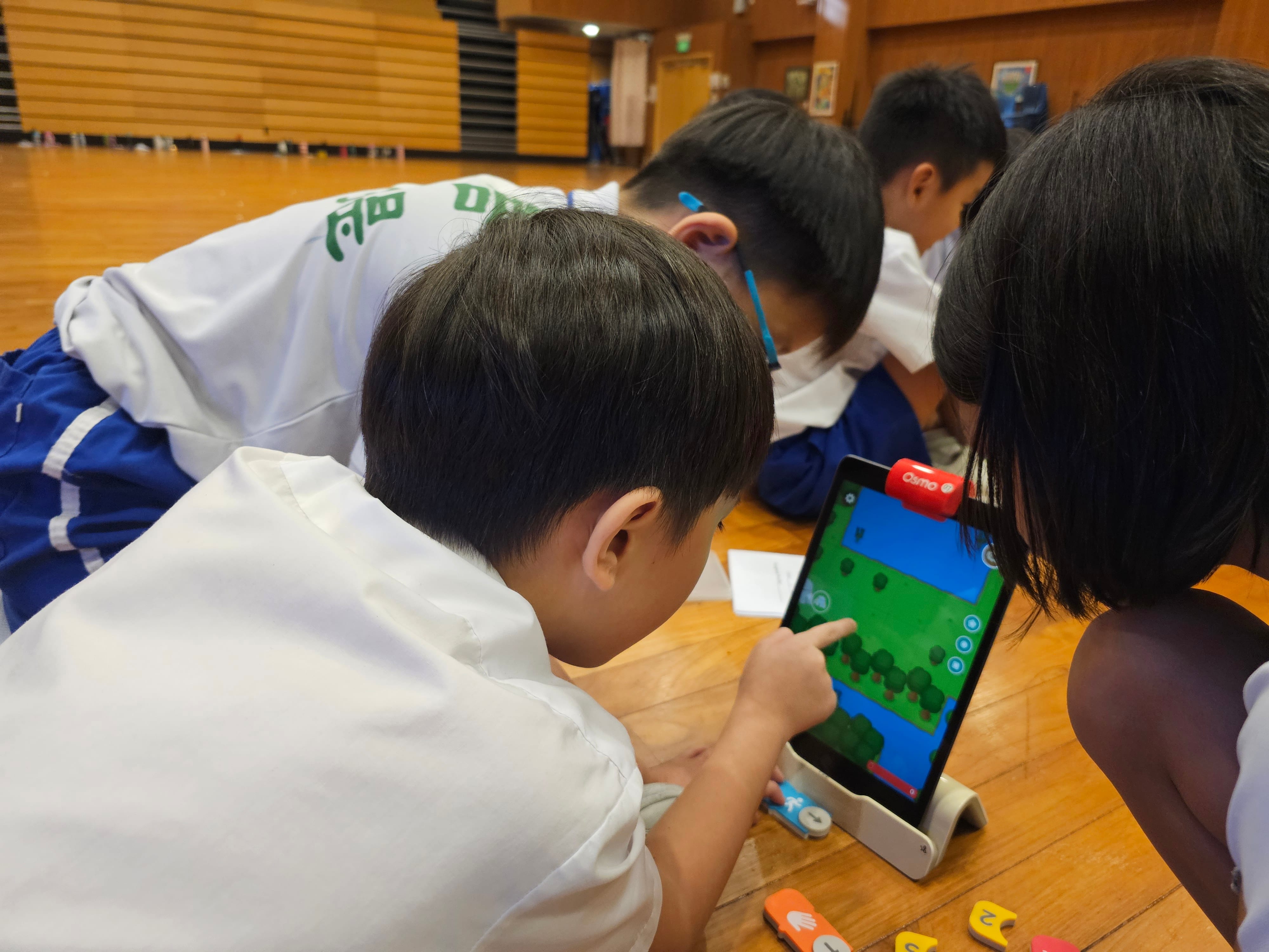 Students in gym uniforms huddle on a wooden floor, one points at a tablet showing a game map.